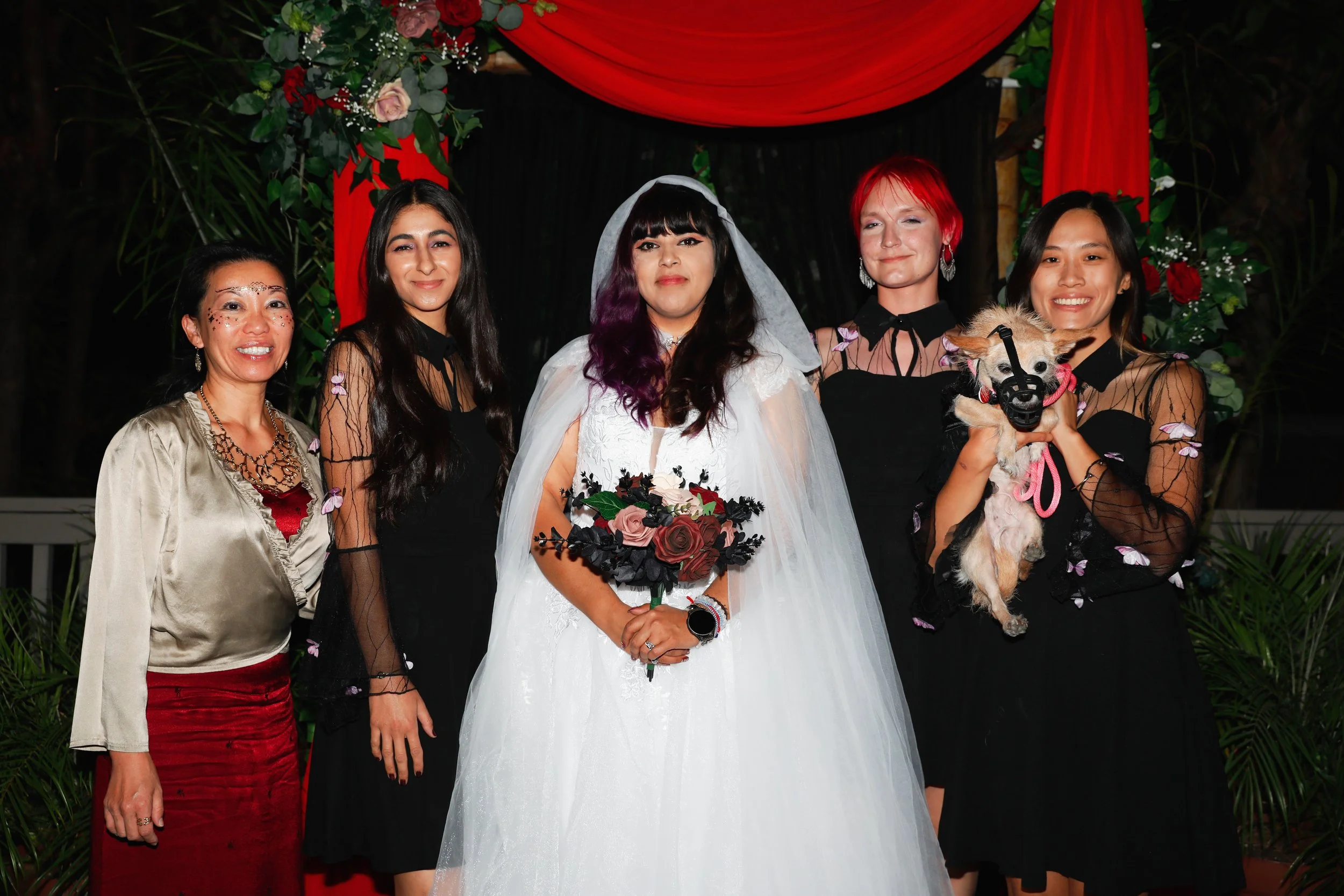 Group of women, with the bride in a white wedding gown holding a bouquet, standing in front of a decorated backdrop, including a woman holding a small dog with a harness, at a wedding celebration.