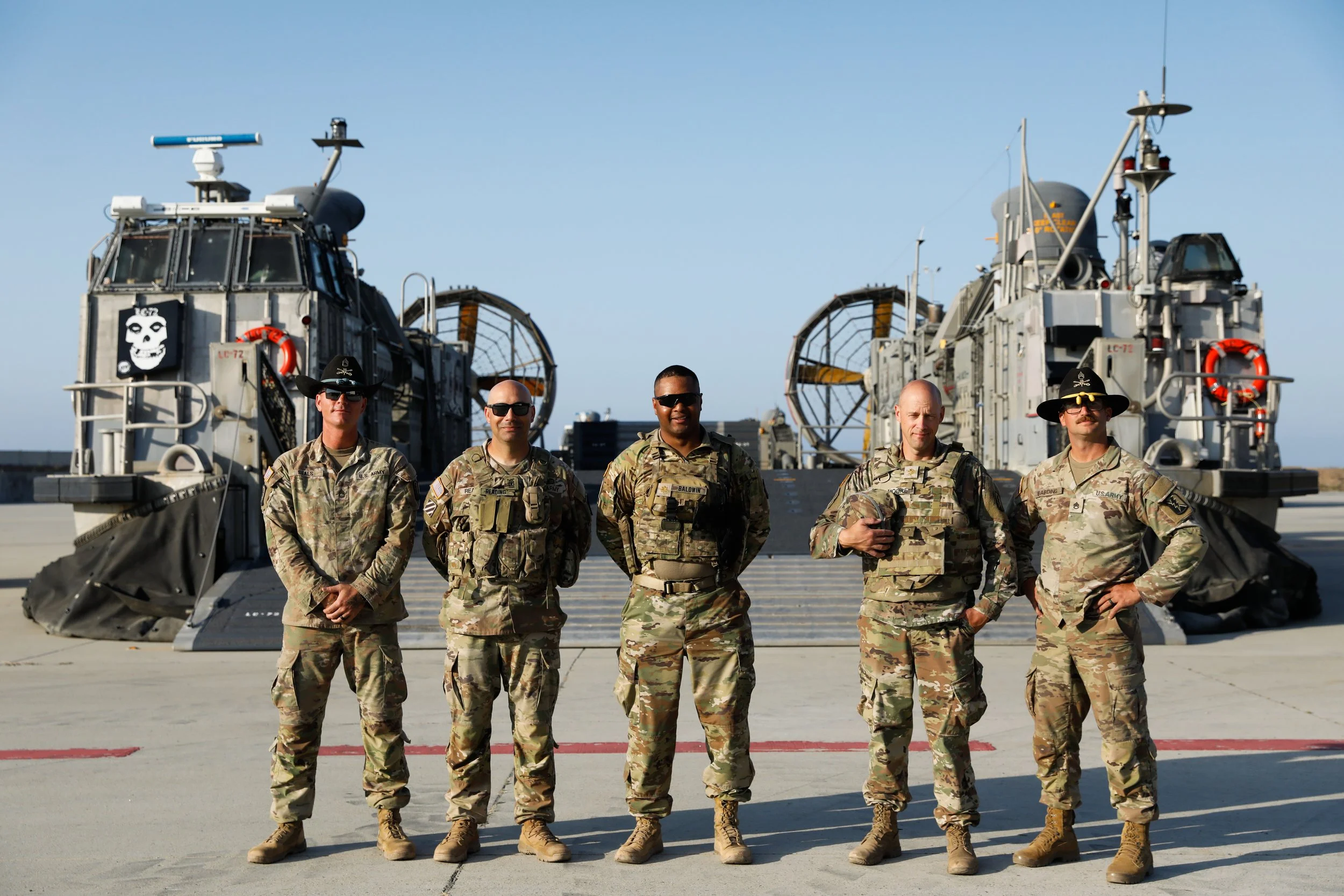 Five soldiers in uniform standing in front of two military aircraft on a tarmac, with a clear sky in the background.