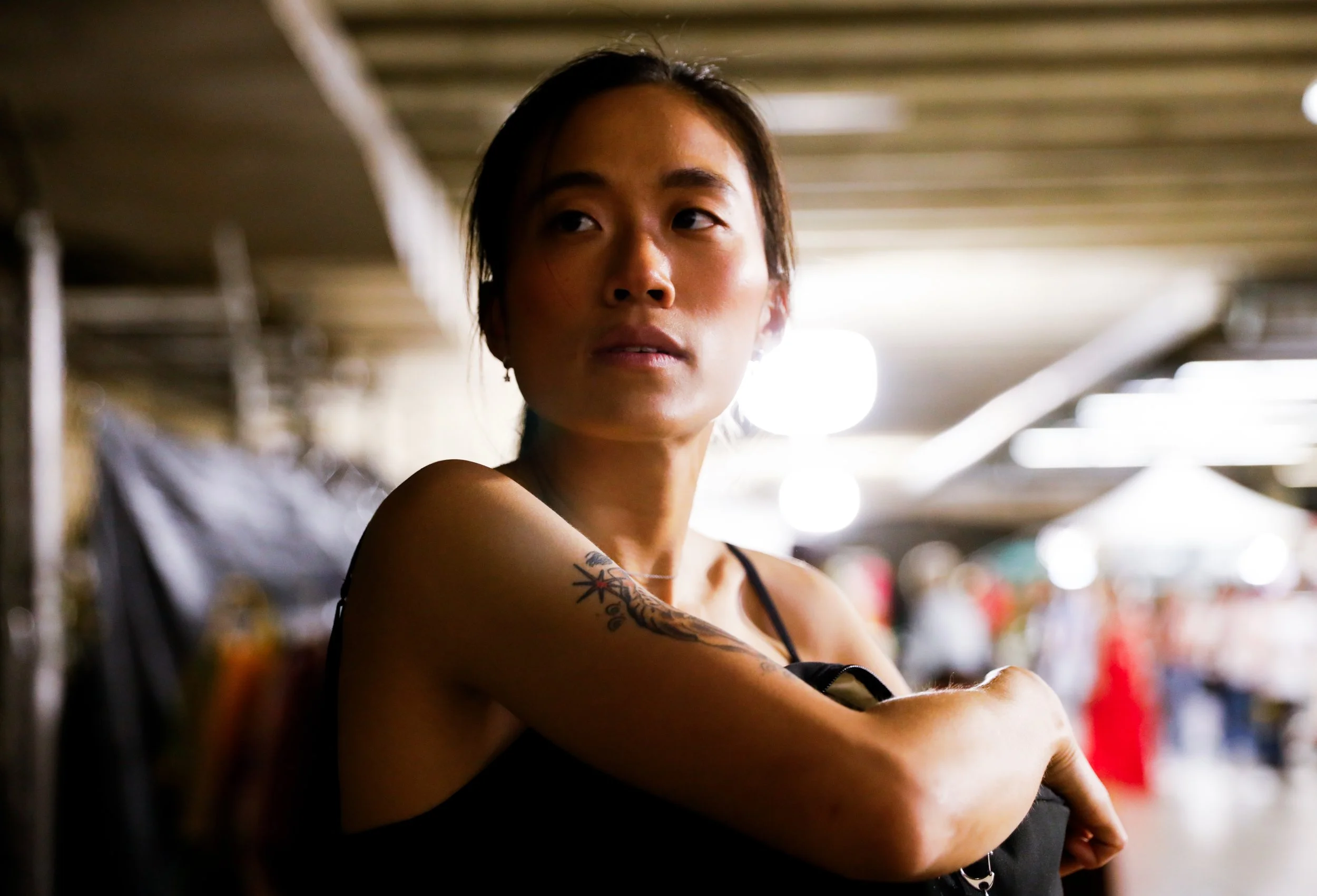 Young woman with shoulder tattoo in a black tank top looking to the side in an indoor market or mall setting.