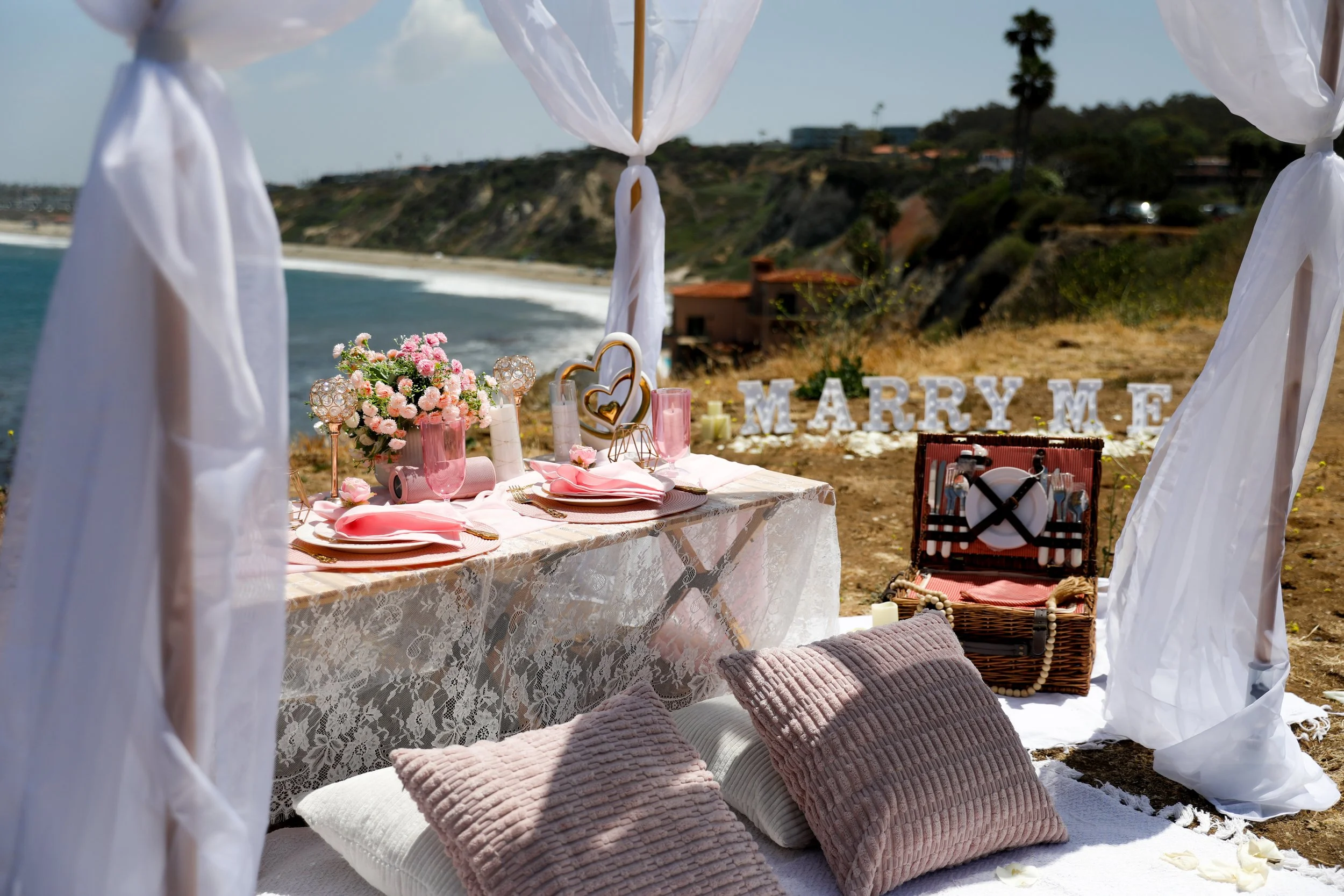 A romantic beachside picnic setup with a table decorated with pink and white flowers, pink candles, and pink dinnerware, framed by sheer white curtains, on a sandy area near the ocean with cliffs and greenery in the background. Cushions are arranged on the ground, and a picnic basket with utensils and a