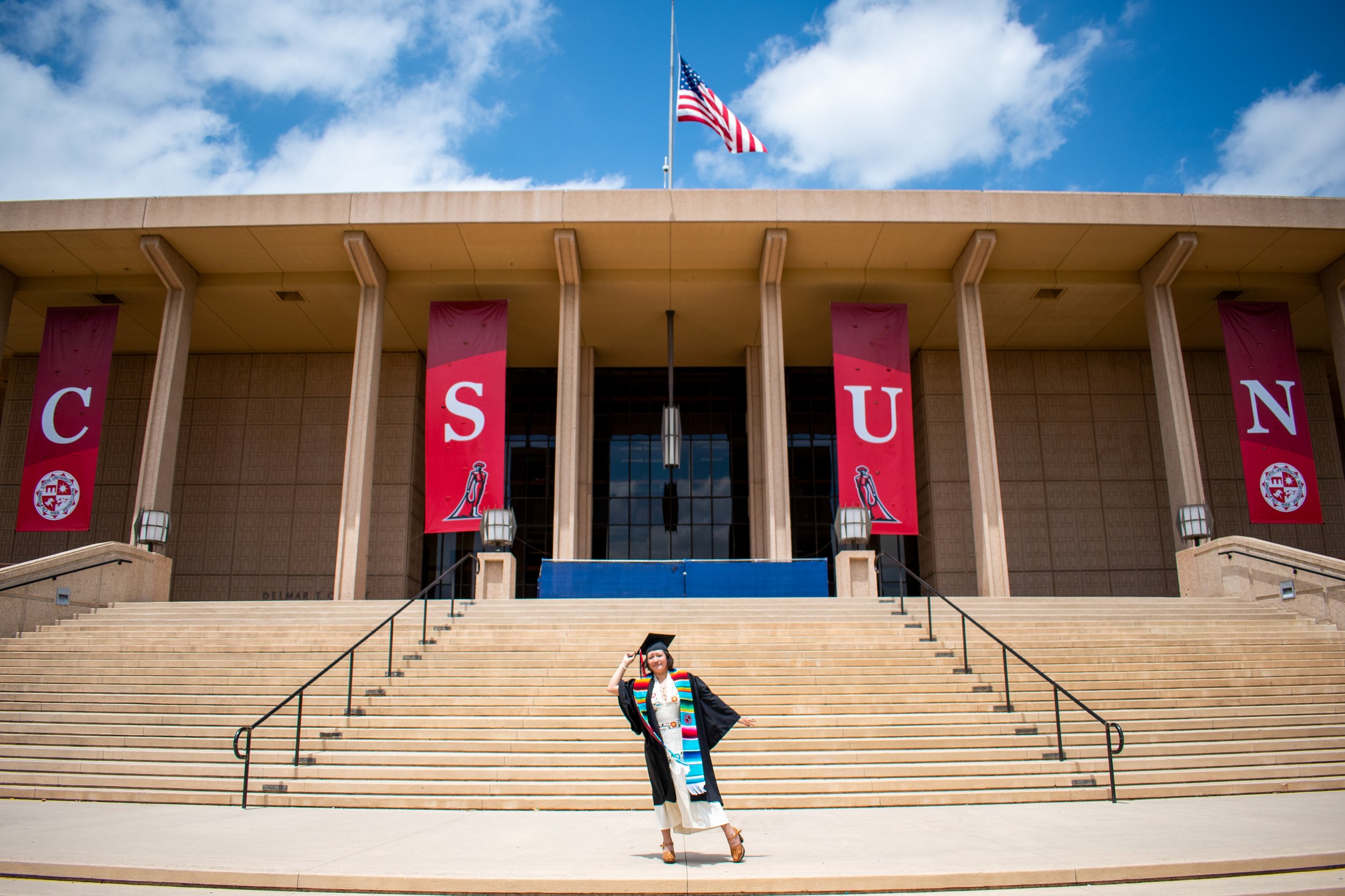 Person in a graduation gown and cap, wearing a colorful stole, standing on steps in front of a large building with banners spelling 'CSUN', with the American flag flying above.