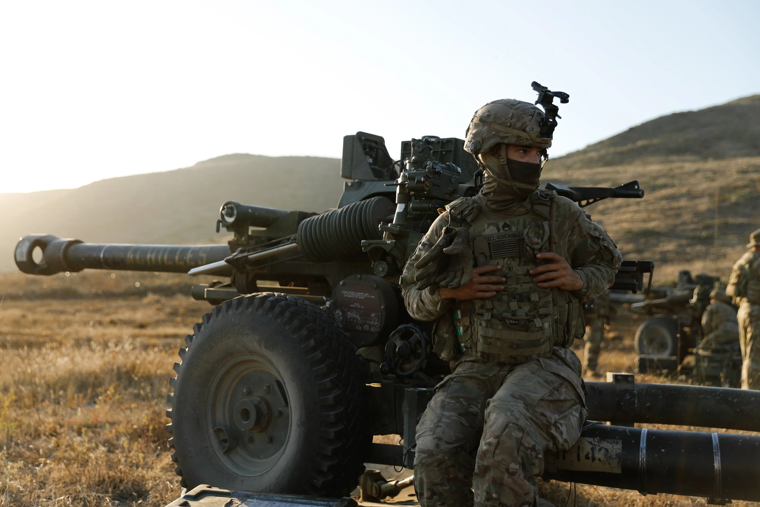 A soldier in camouflage uniform and a helmet sitting on a large military artillery gun in a field with hills in the background at sunset.