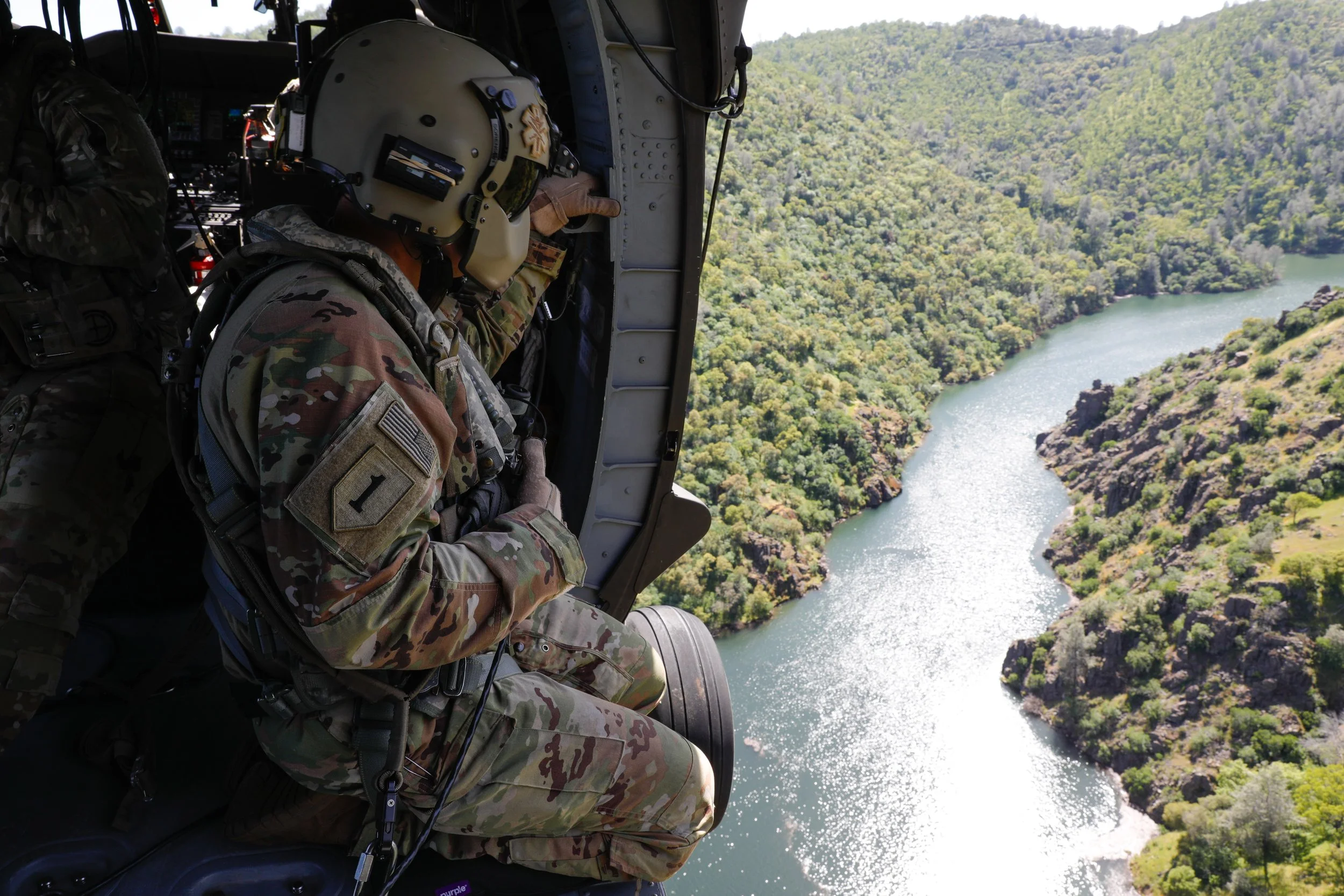 Military personnel in camouflage uniforms and helmets sit inside a helicopter flying over a river canyon with lush green trees on both sides.