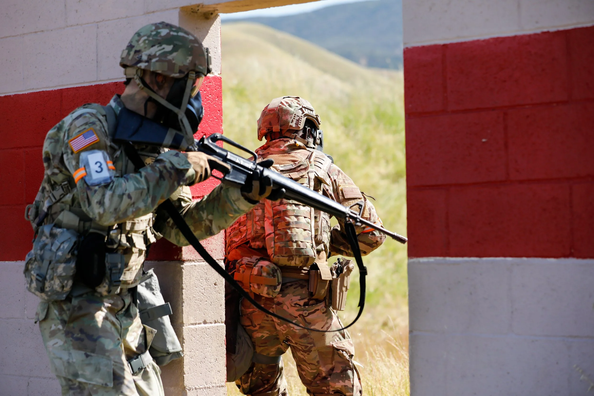 Two soldiers in camouflage uniforms and helmets taking cover behind a wall with red and white stripes during a military exercise.