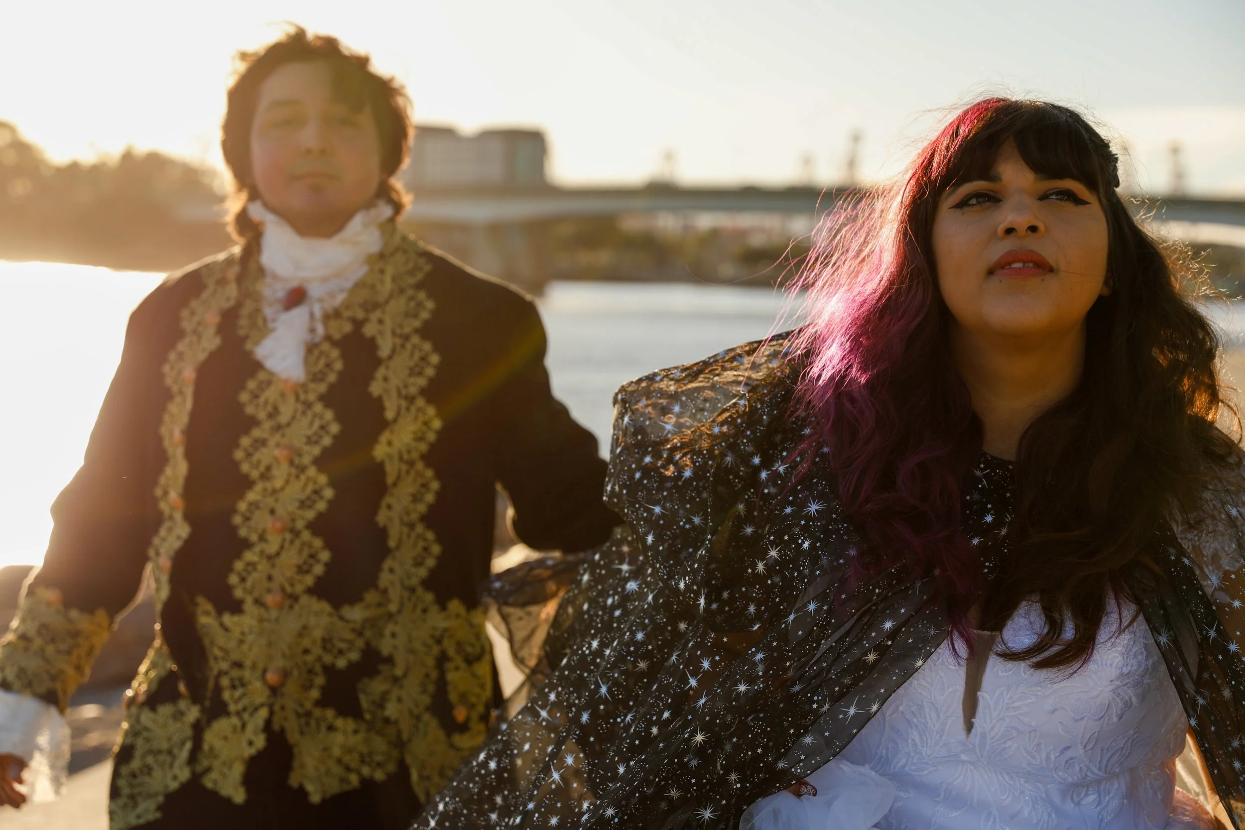 Two individuals dressed in elaborate costumes walk on a beach at sunset, with a bridge spanning a body of water in the background.