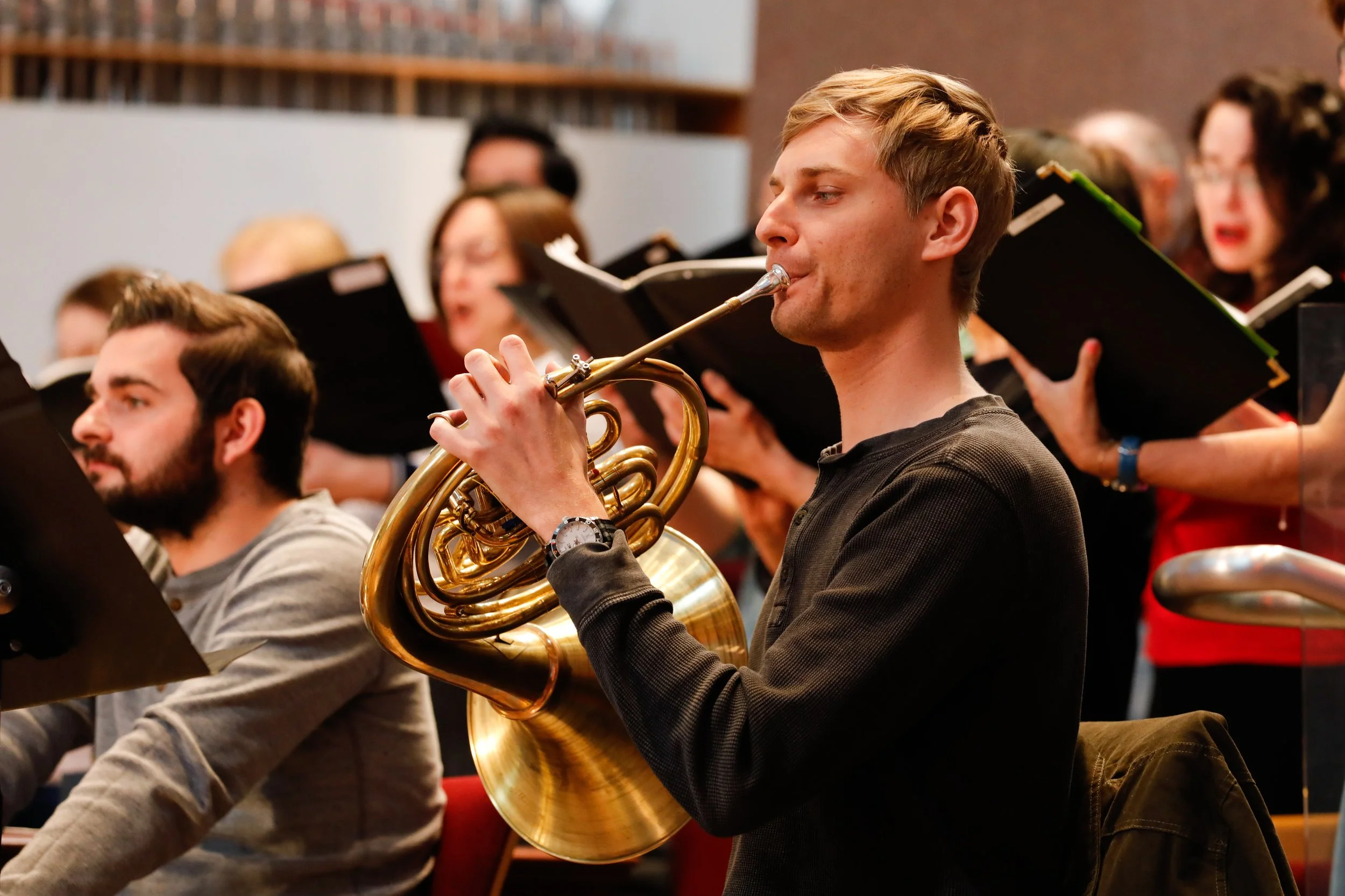 A young man playing a French horn during a choir rehearsal with singers holding sheet music in the background.
