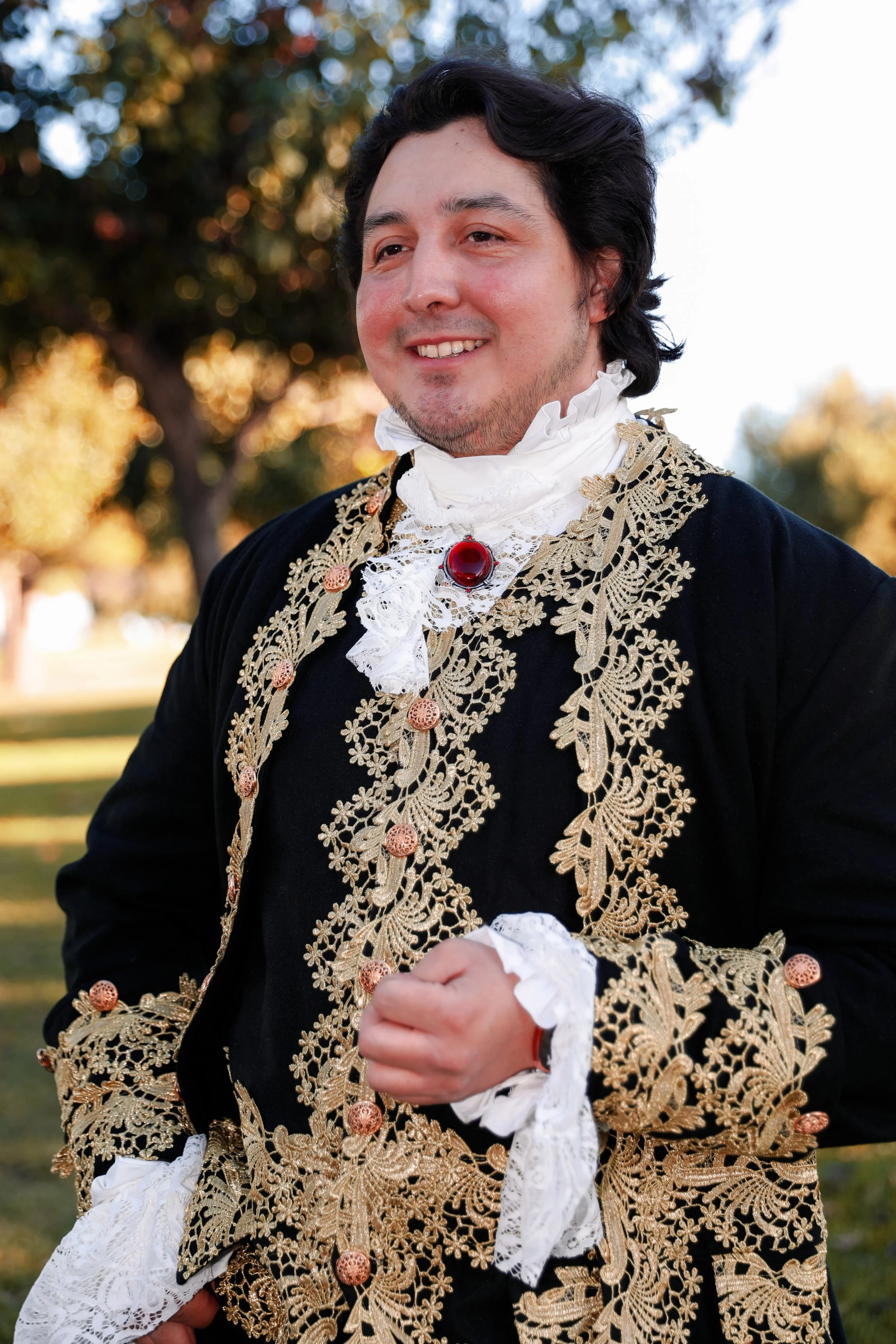 A man dressed in a flamboyant historical costume with black velvet fabric, elaborate gold embroidery, lace cuffs, and a red gemstone brooch, standing outdoors with trees in the background.