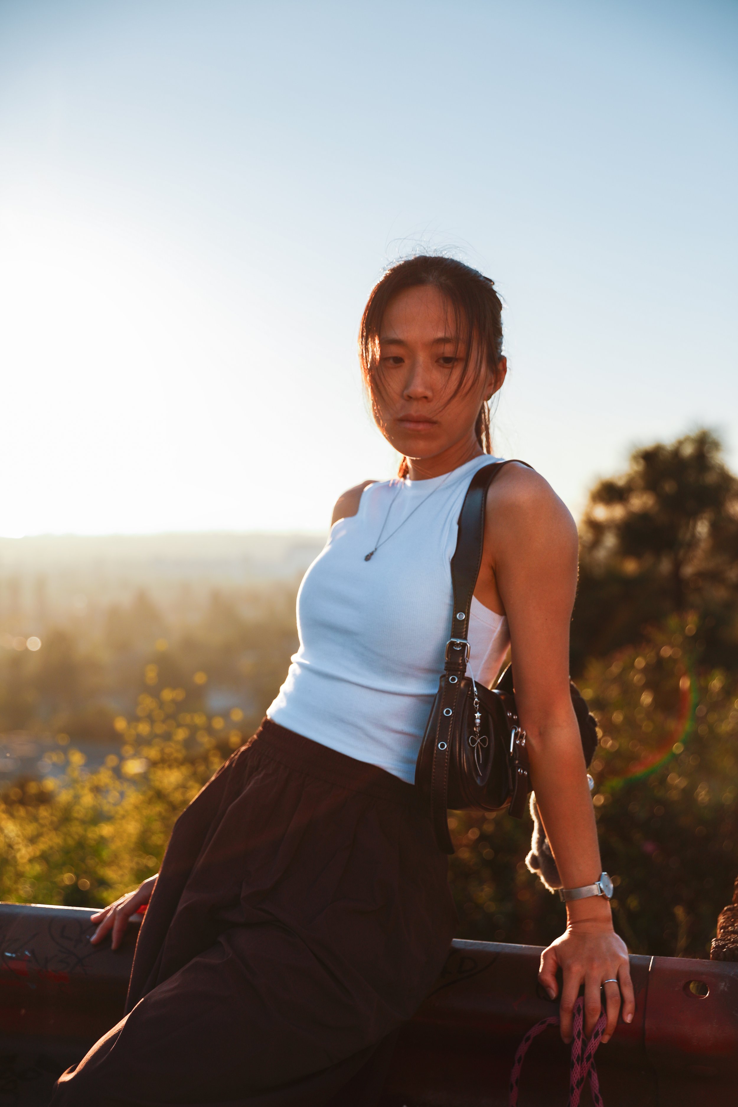 A young woman in a white sleeveless top and black pants leaning on a railing outdoors during sunset, with blurred trees and a clear sky in the background.