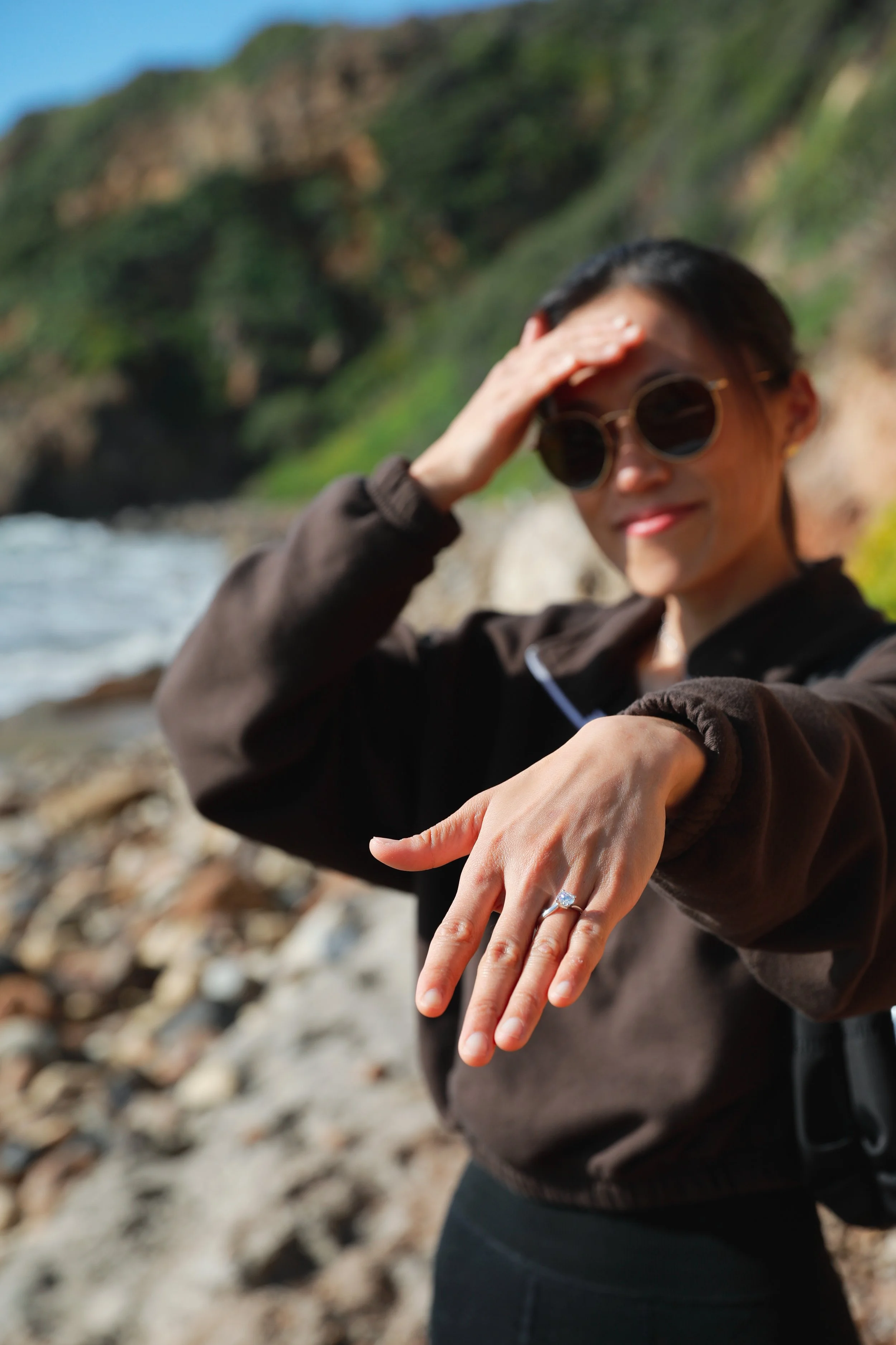 A woman outdoors near a rocky riverbank, wearing sunglasses and a brown jacket, showing her diamond engagement ring on her finger.