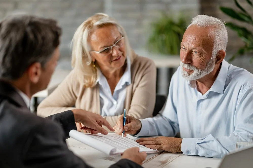 Three elderly adults, two men and one woman, sitting at a table during a discussion or consultation, with one man holding a pen and a document.