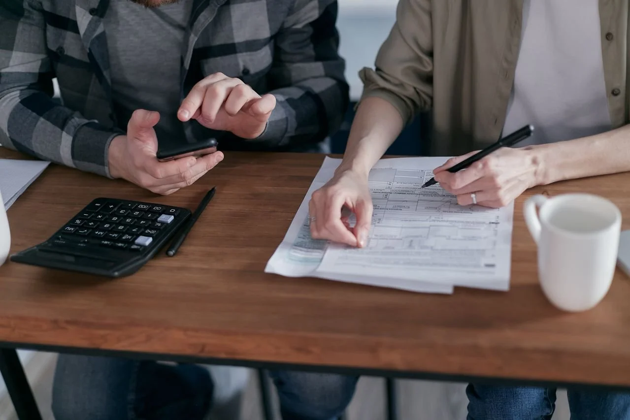 Two people review documents at a wooden table, with one using a pen and the other holding a smartphone, amid papers, a calculator, and a coffee mug.