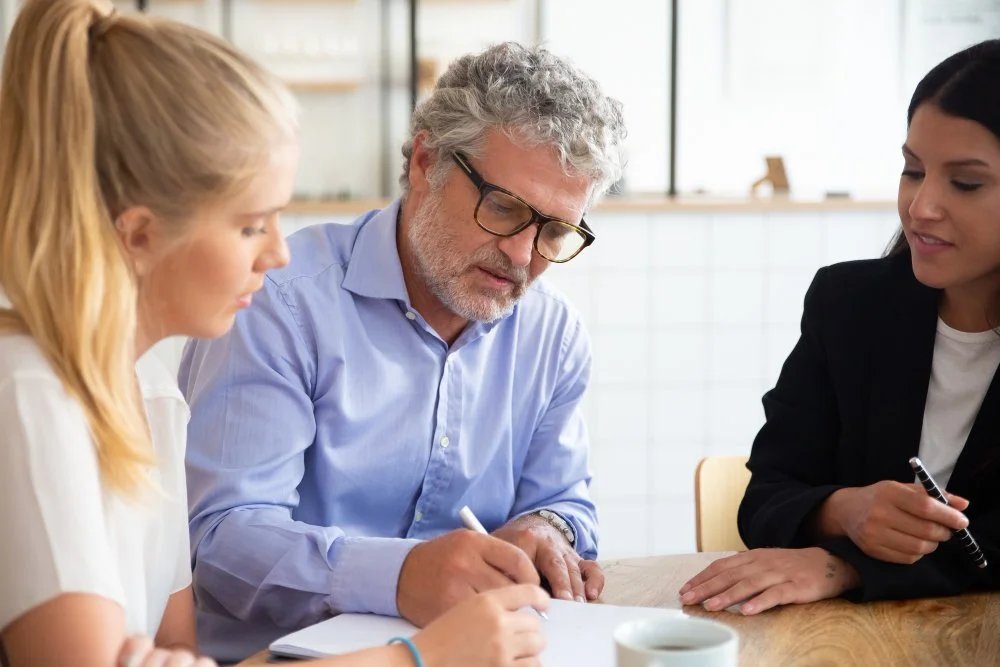 Three people sitting at a table in a bright office, engaged in a discussion or meeting, with a woman on the left taking notes.