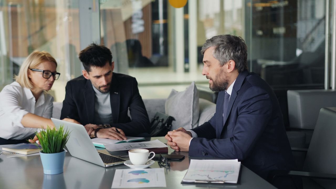 Three professionals in a business meeting, two women and one man, sitting around a table with documents, a laptop, and a coffee cup, engaged in discussion.