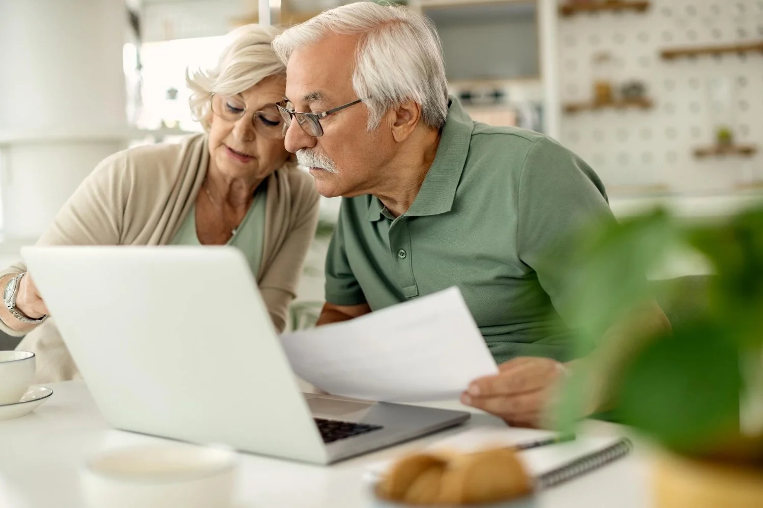 An elderly couple reviews documents together at a desk with a laptop in front of them, in a bright kitchen or dining area.