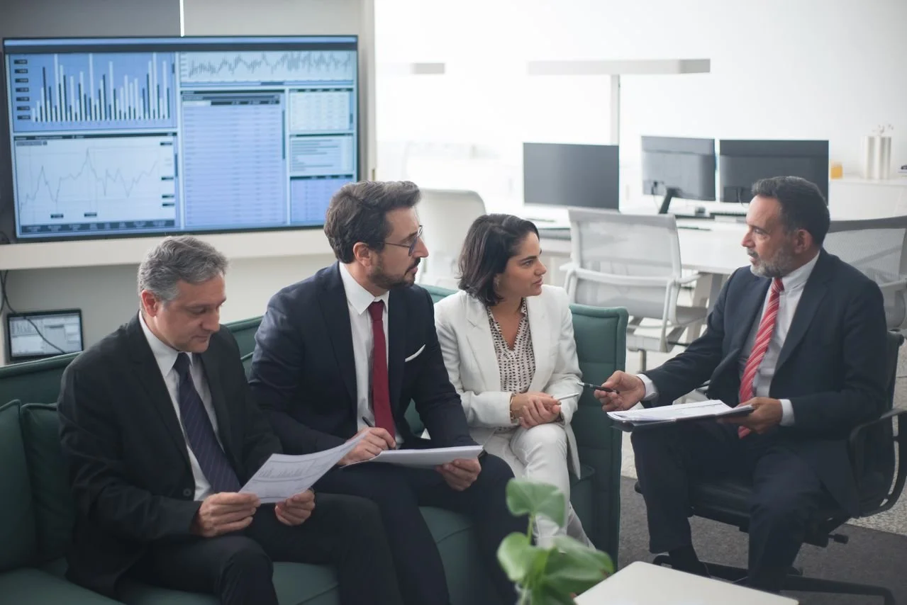 Four professionals in business attire sitting on sofas and engaging in a discussion in a modern office, with multiple computer monitors and financial data displayed on a large screen in the background.