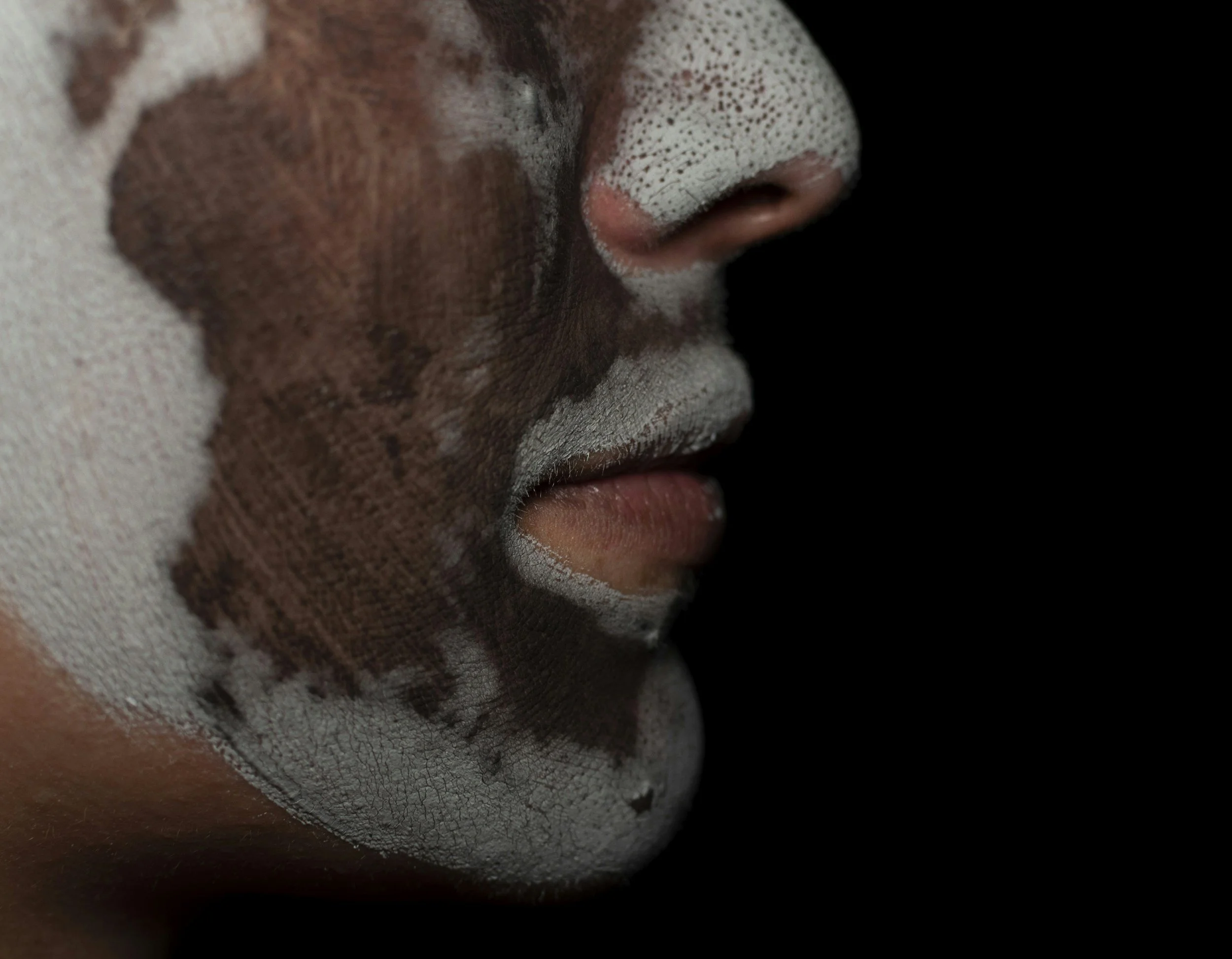 Close-up of a person's face with white and brown face paint, showing the nose, lips, and a portion of the cheek against a black background.