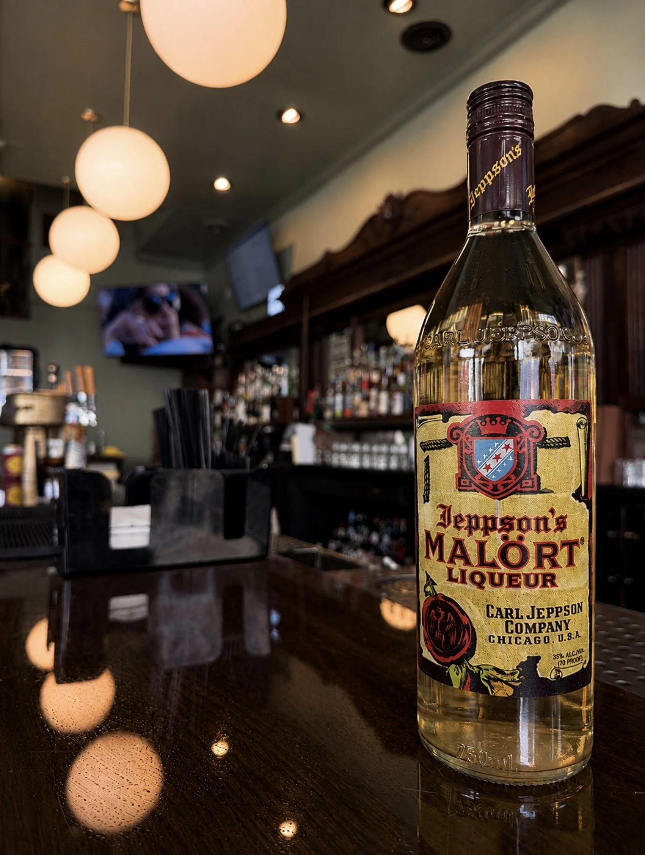 A bottle of Jeppson's Malört Liqueur on a bar counter inside a dimly lit bar or restaurant. The background features hanging round lights and a television screen. The bar has various bottles and bar equipment.