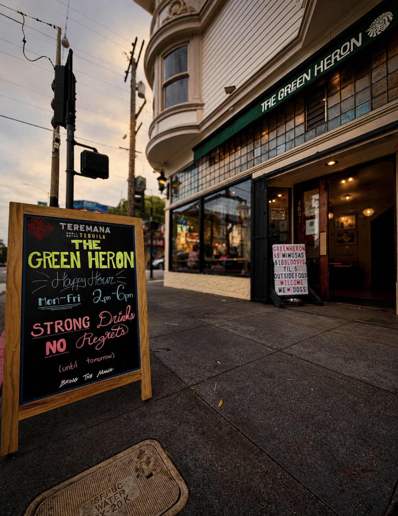 A sidewalk scene outside a bar called "The Green Heron" featuring a chalkboard sign with happy hour details, a smaller sign with food and drink offers, and a building with large windows and a green awning. The sky is cloudy, and there are utility pol
