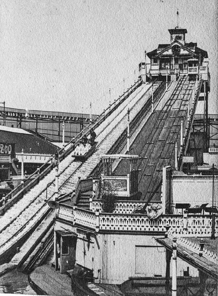 Black and white photograph of a historic amusement park ride with a sloped track, possibly a roller coaster or funfair slide, on a boat-like structure with decorative railings and a small building at the top.