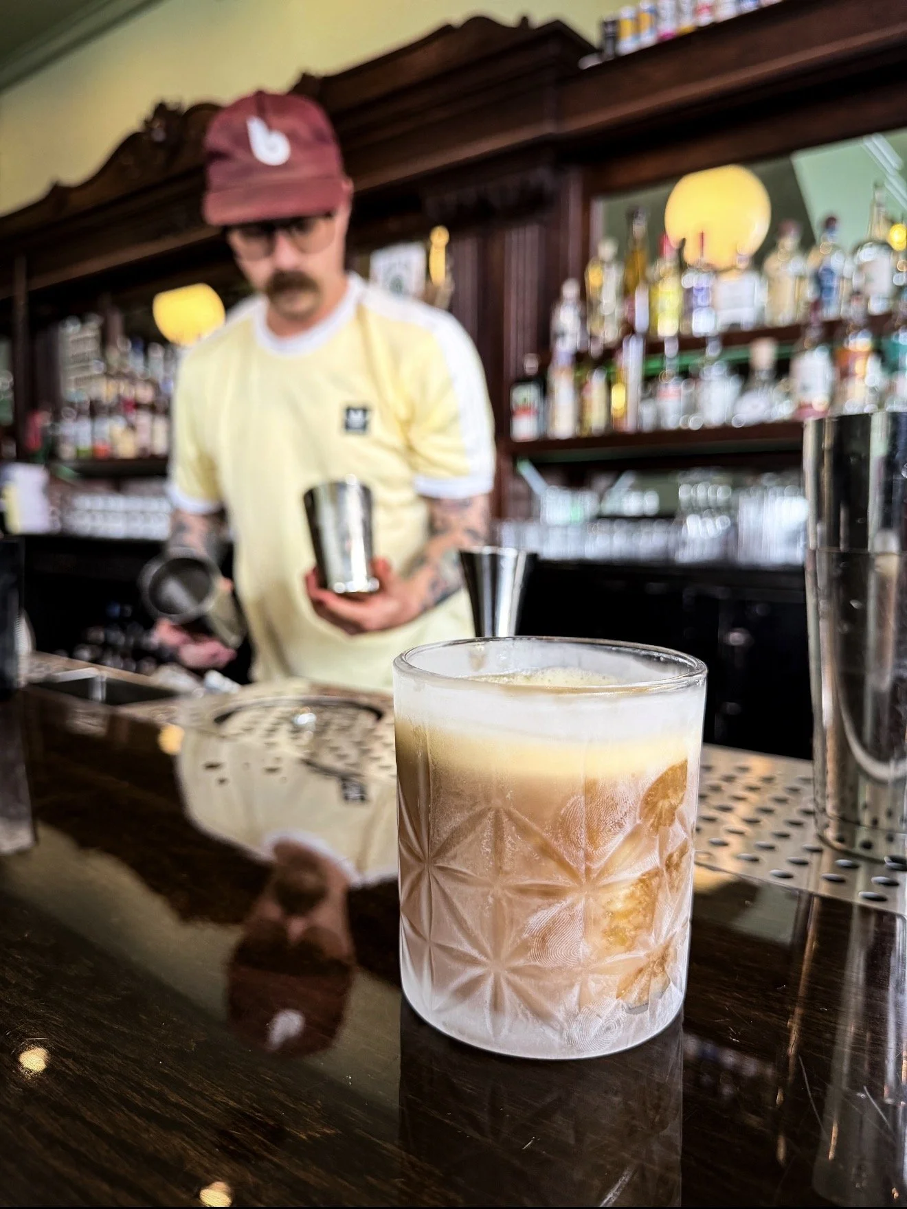 A close-up of a glass filled with a layered cocktail with ice, in the foreground, on a bar counter. In the blurry background, a man with a mustache, glasses, a maroon cap, and a yellow shirt is preparing drinks behind a bar with shelves of bottles.