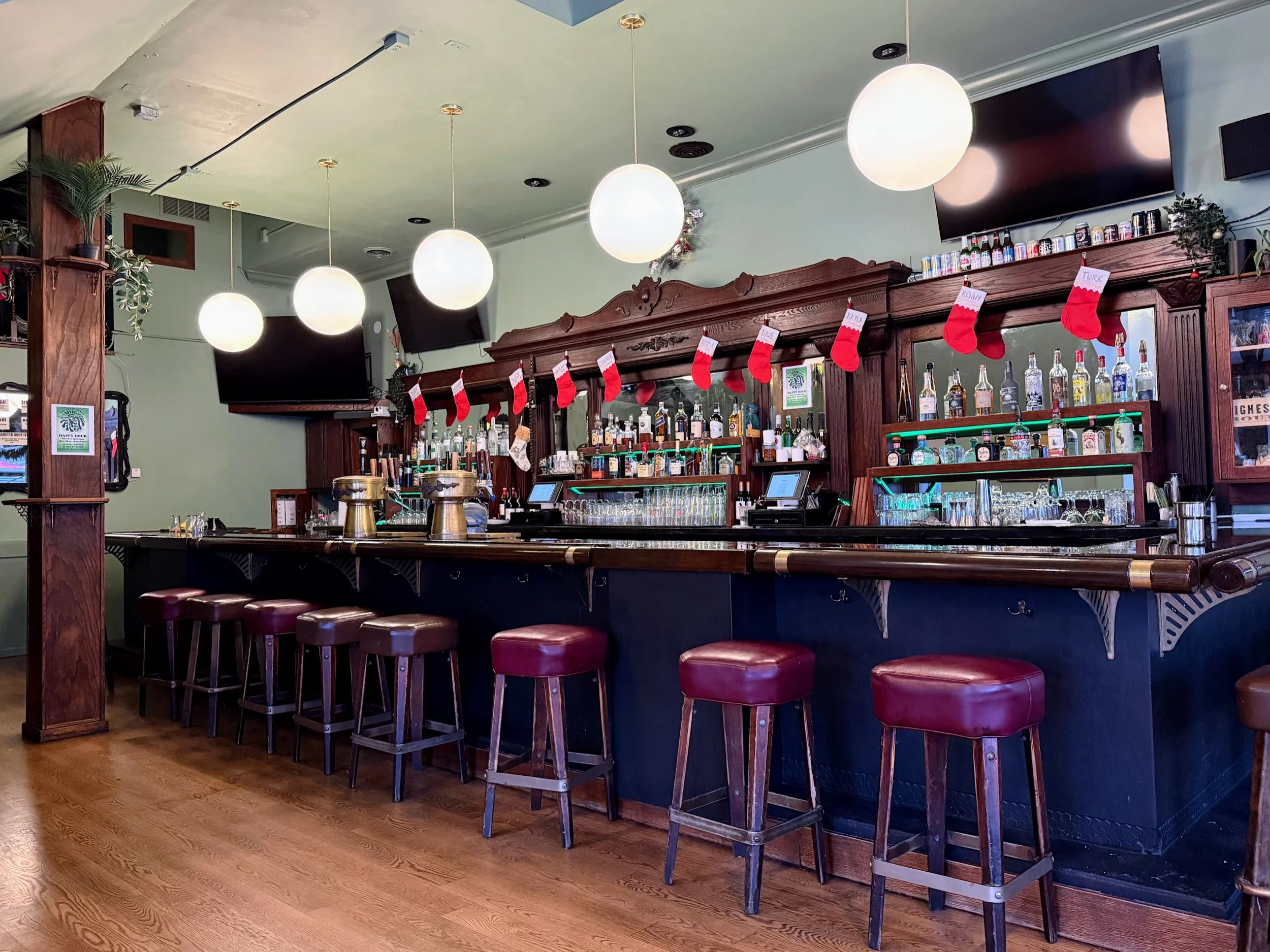 Interior of a bar with a wooden counter, red bar stools, and Christmas stockings hanging from the shelf behind the bar, decorated for the holiday season.