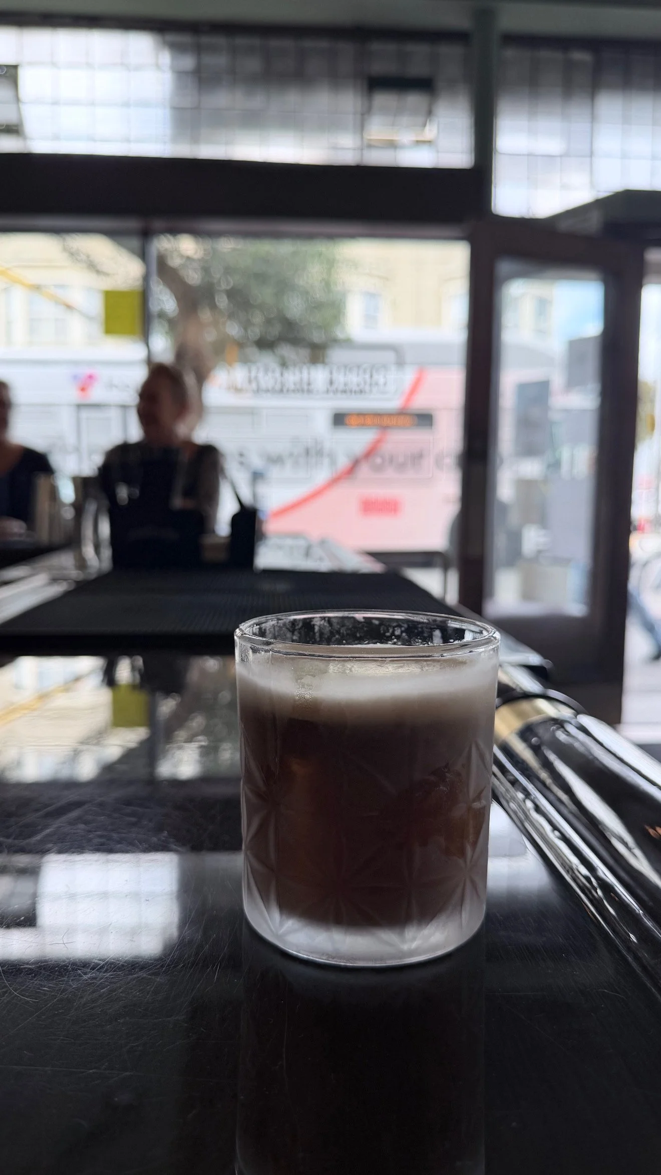 Close-up of a glass of layered coffee or mocha on a table, with a blurred café interior and people in the background.