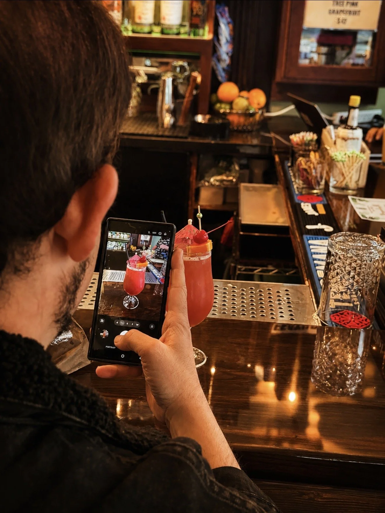 A person taking a photo of a pink cocktail with cherry and umbrella on a bar counter.