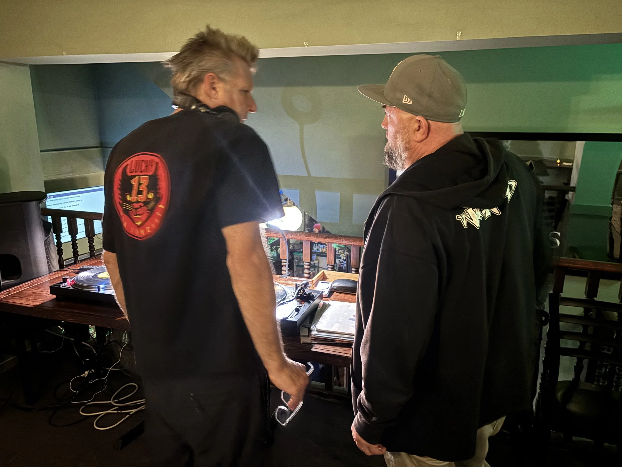 Two men standing face to face in a room with DJ equipment, including turntables, in the foreground. One man is wearing a black sweatshirt with a logo on the back, and the other is wearing a hoodie and cap. They are engaging in conversation or interac