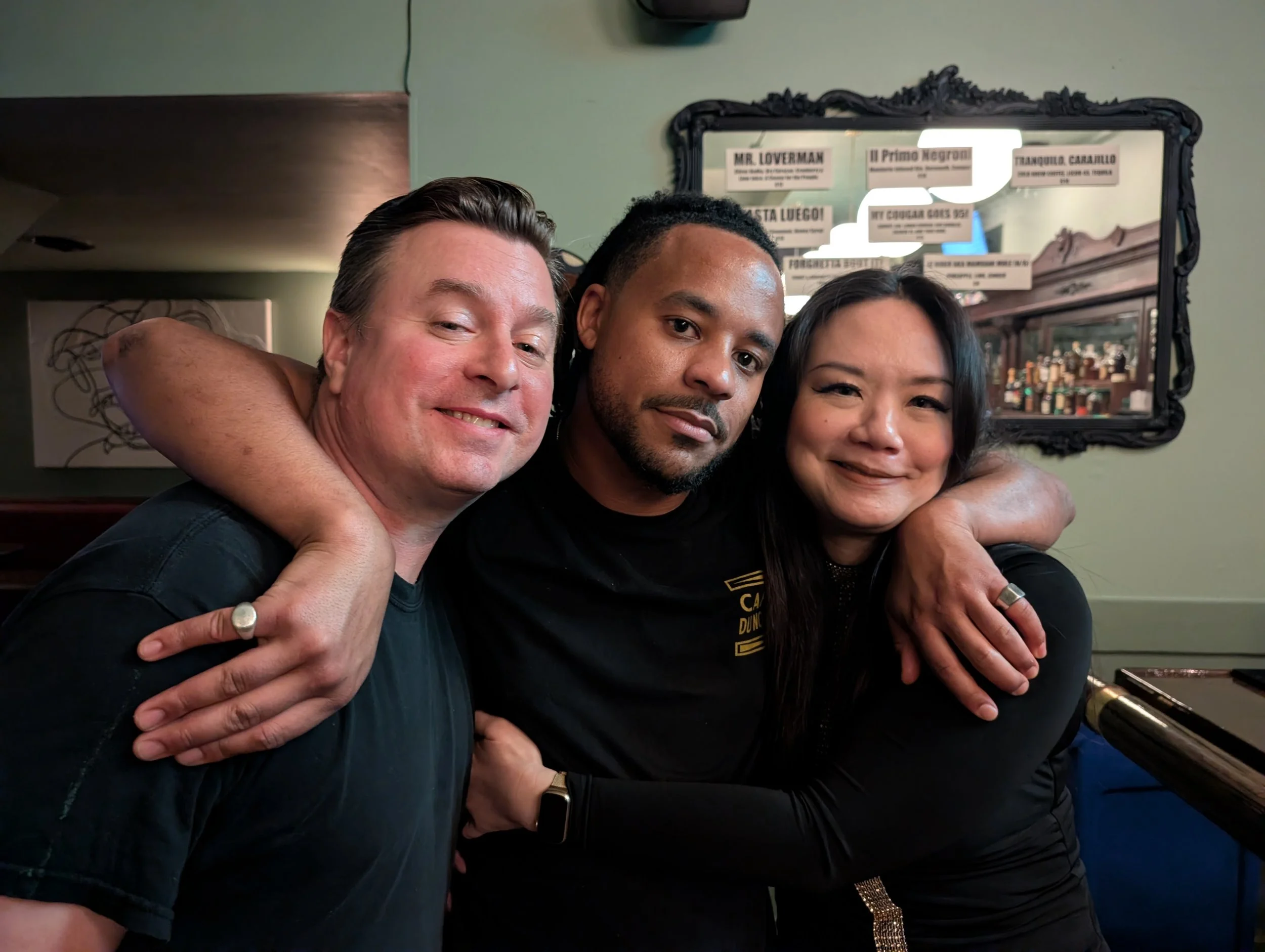 Three people hugging and smiling in a cozy restaurant or bar, with a mirror and signs in the background.
