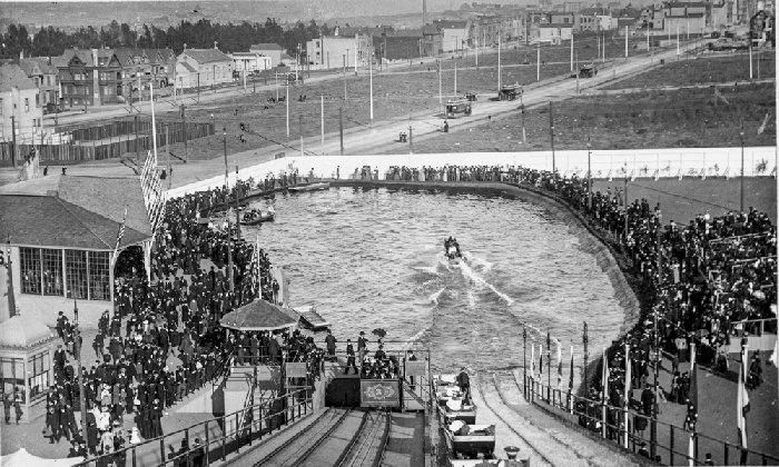 Crowds line a curved man-made lake as a boat speeds across the water on the Haight Street Chutes opening day in 1895.
