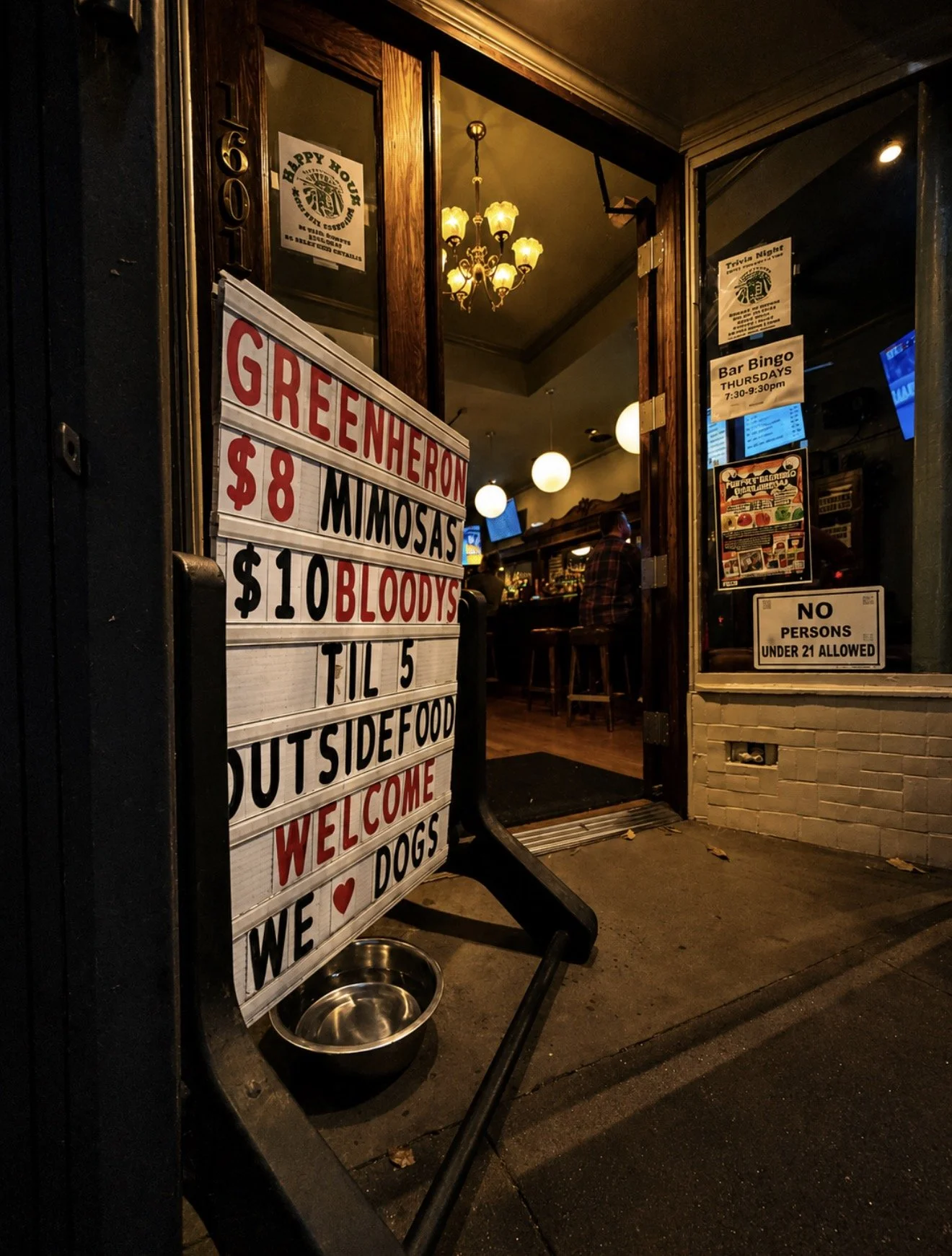 The image shows the entrance of a bar or restaurant with a sign listing food and drink specials, including Green Hero, $8 Mimosas, $10 Bloody Marys, and outdoor dining hours. Inside, visible through the door, are hanging lights and a person sitting a