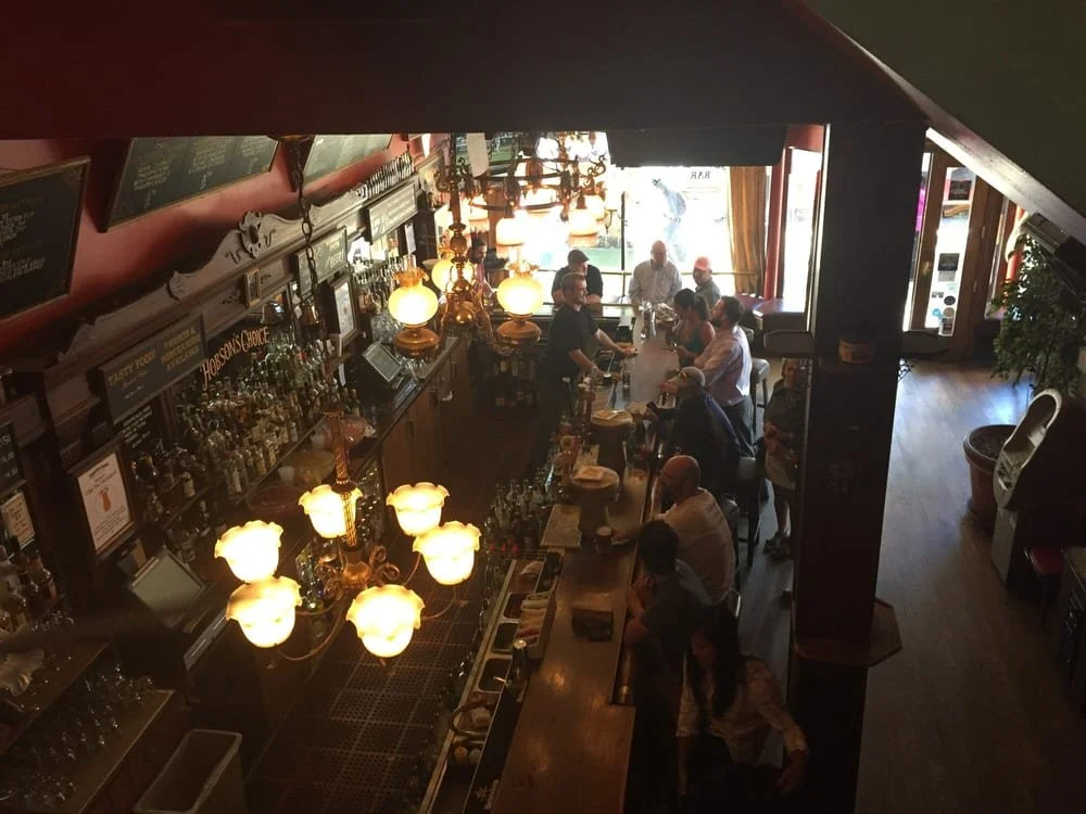 The Hobson&rsquo;s Choice bar seen from the upstairs lounge, with antique gas-lamp chandeliers, a long back bar of bottles, and patrons gathered below.