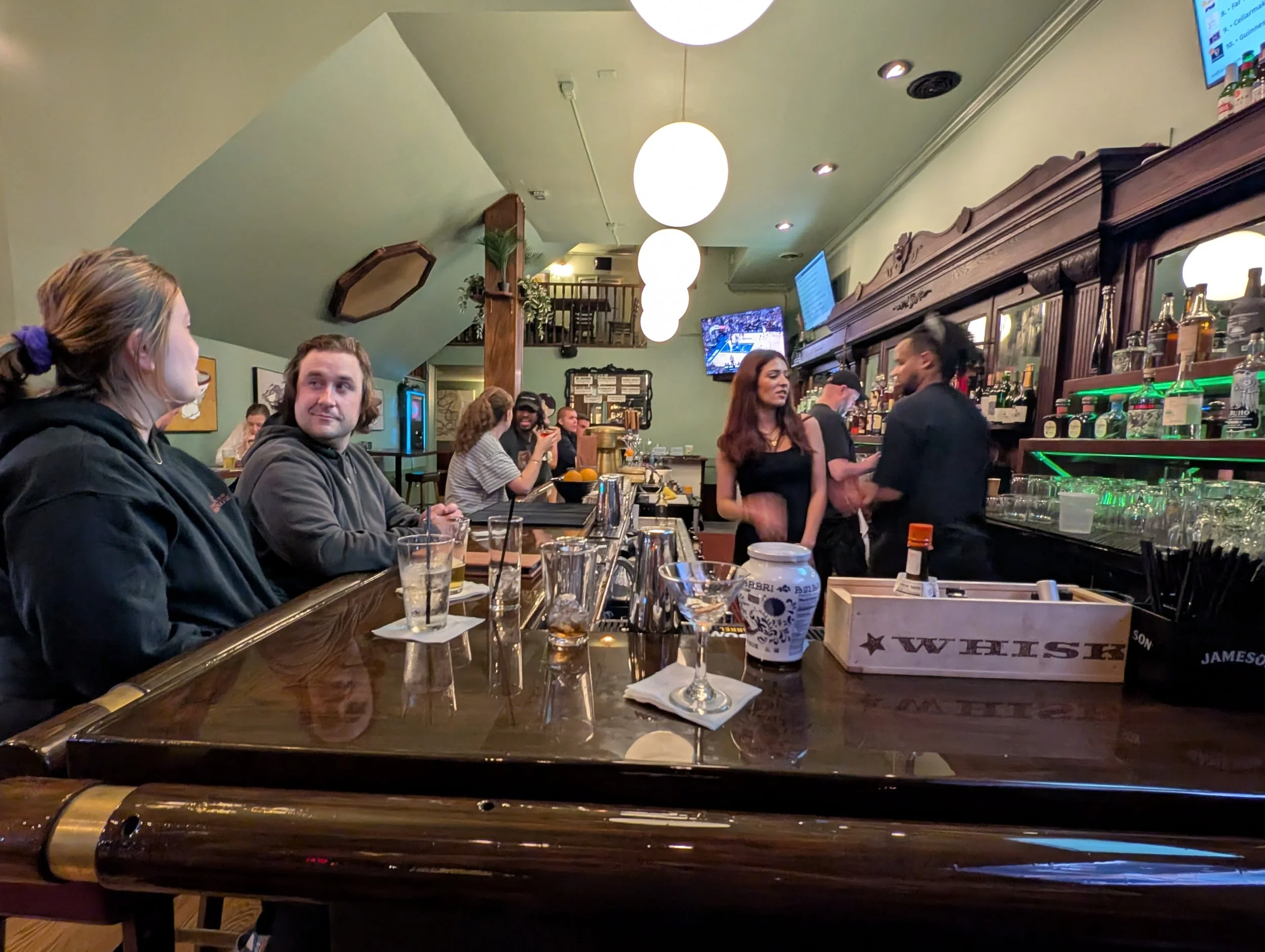 Inside a lively bar or restaurant with patrons seated at the bar and standing, engaging in conversation. There are endless bottles of liquor behind the bar, illuminated by green lighting. Overhead lighting consists of large, spherical white fixtures.