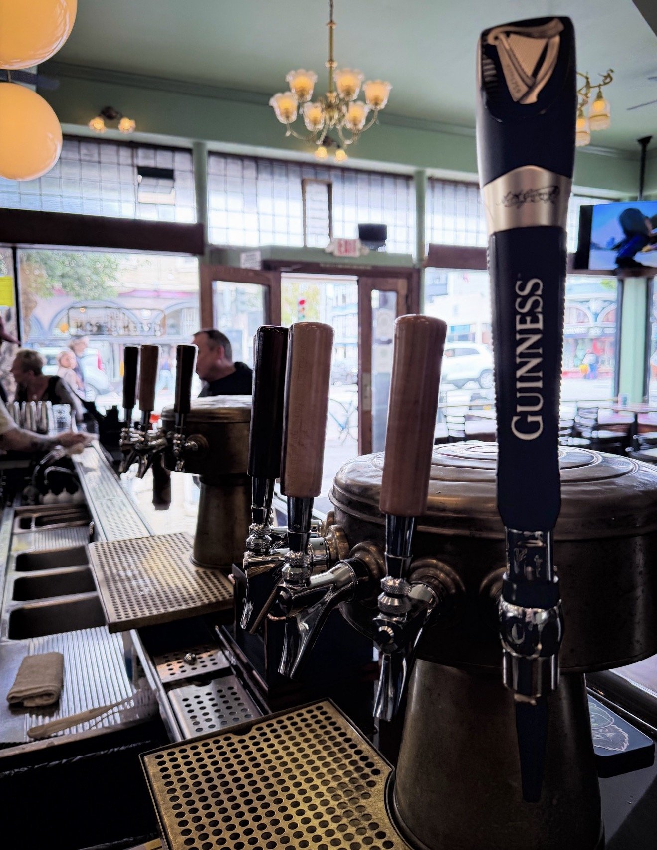 Close-up of Guinness tap handles at a bar with people sitting in the background and large windows letting in natural light.