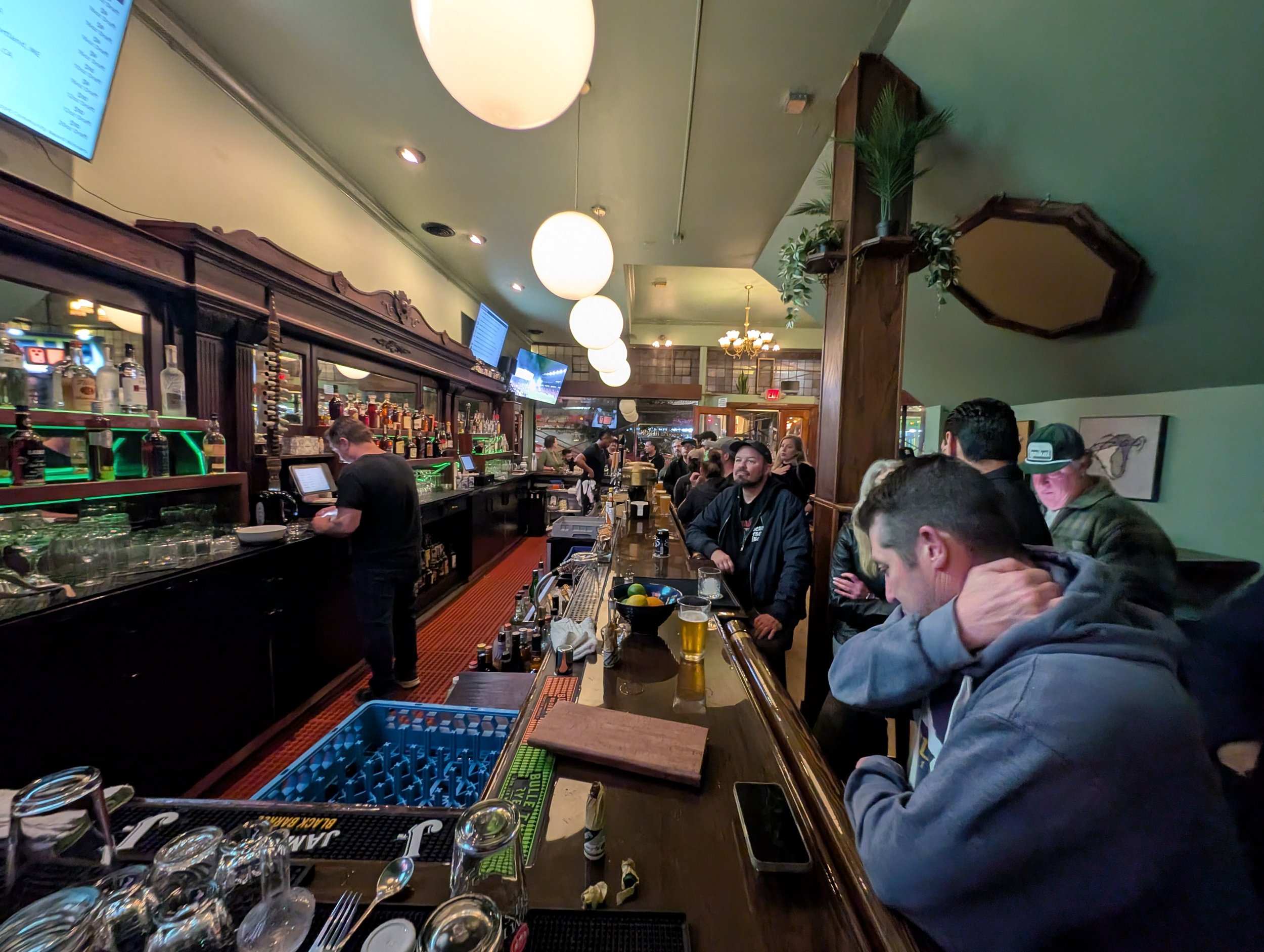 A busy bar with people sitting and standing, drinks on the counter, and bartender working. Round ceiling lights and TVs are visible in the background.