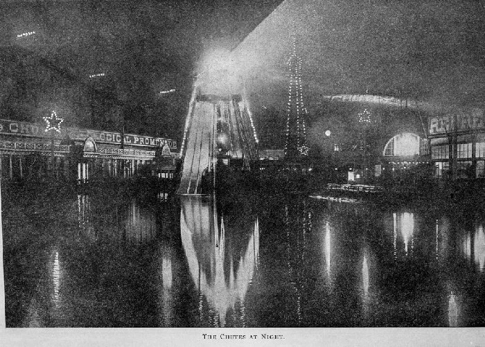 Black and white photo of the miniature Ferris wheel at nighttime, lit up and reflected in a water surface, with carnival buildings in the background.