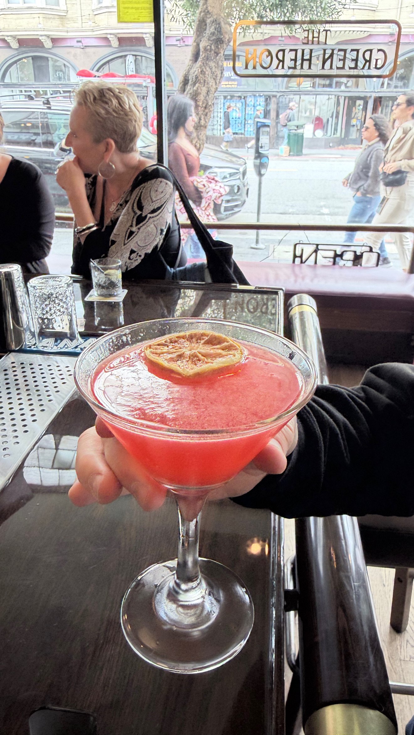 View from inside a restaurant showing a person holding a pink cocktail with a lemon slice garnish in the foreground. Outside the window, pedestrians walk on the street with buildings and cars visible.