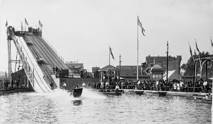 A boat splashes down at the base of the tall wooden water-chute ramp as spectators watch from a pier in 1895.