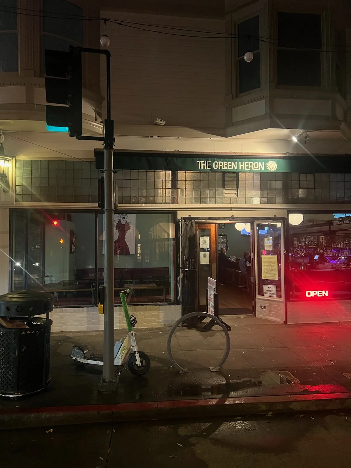 Nighttime street scene featuring a bar called 'The Green Heron' with an open sign, windows showing bar seating, a scooter and bicycle parked outside, and a traffic light with a green pedestrian signal.