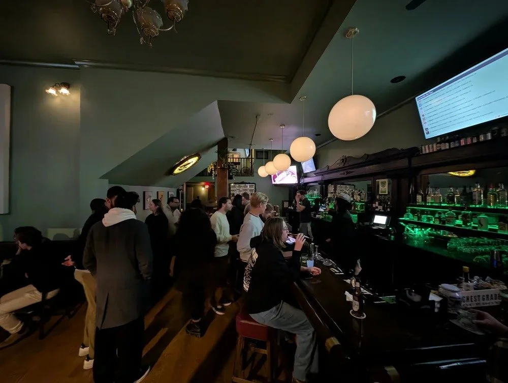 The Green Heron&rsquo;s main room in December 2025: warm globe pendant lights, a long dark bar backlit in green, and patrons gathered beneath the high ceiling.