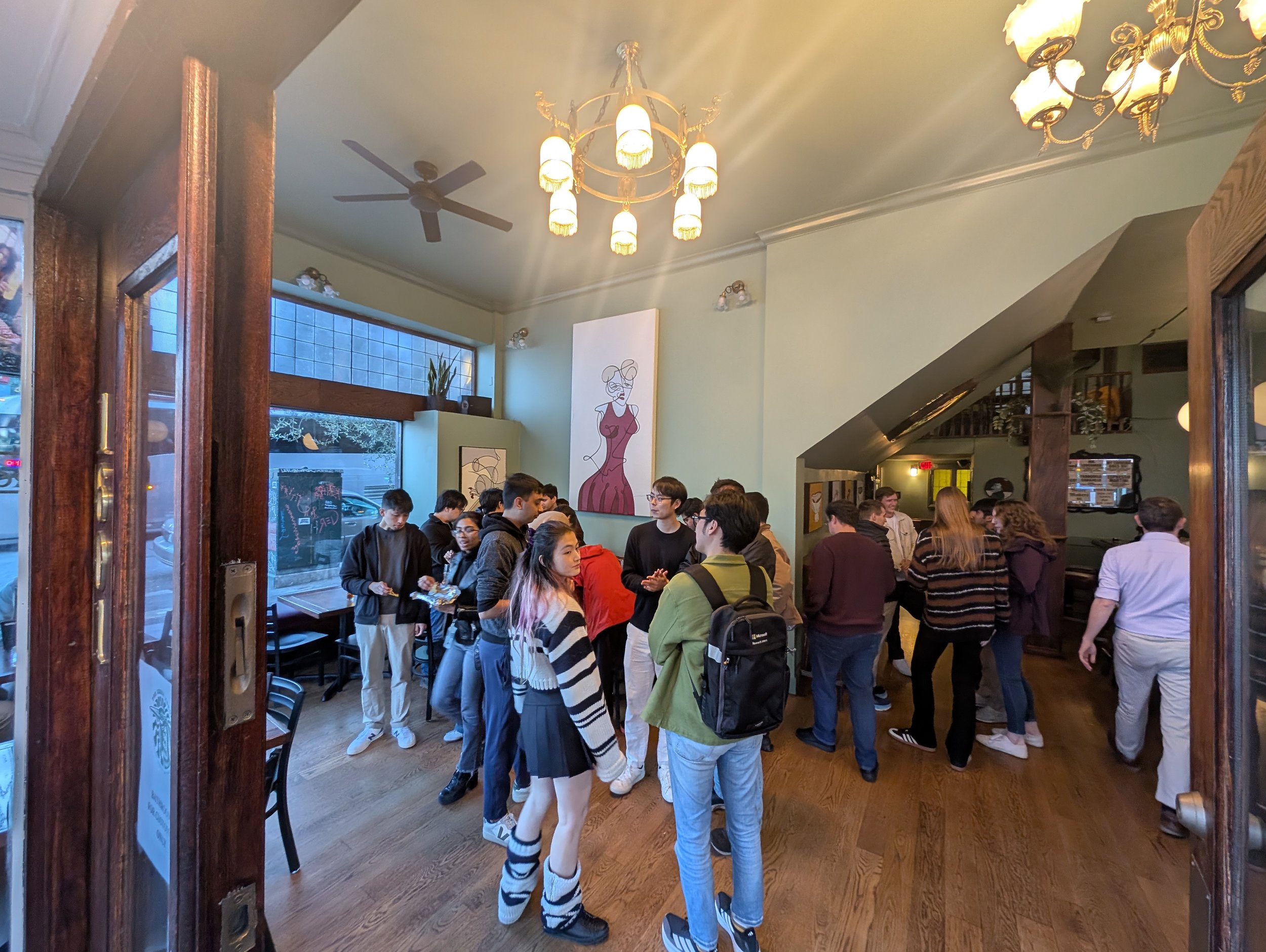 A group of people standing and talking inside a restaurant with wooden floors, green walls, and hanging light fixtures, seen through a doorway.