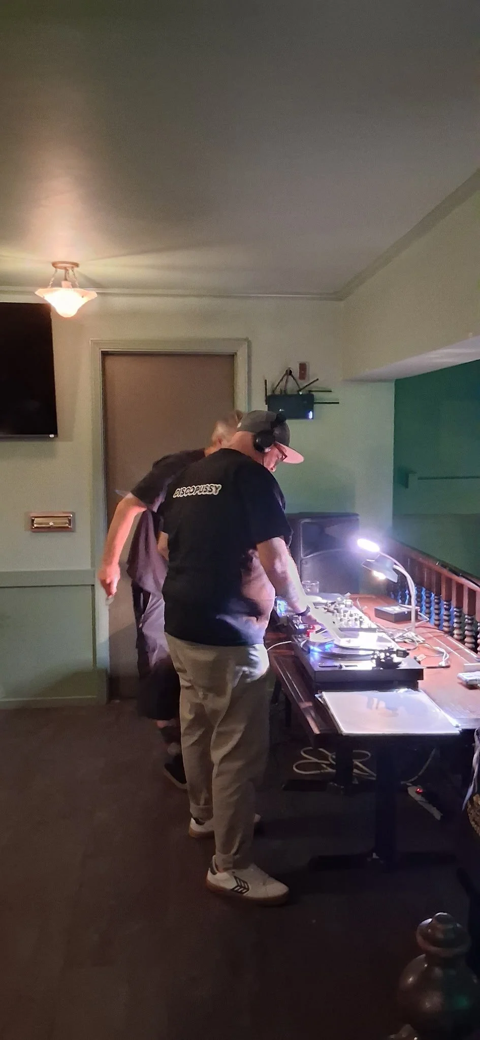 A person wearing a black t-shirt with the word 'DISCOPUSSY' on the back is DJing at a party, standing at a table with DJ equipment in a dimly lit room with green walls.