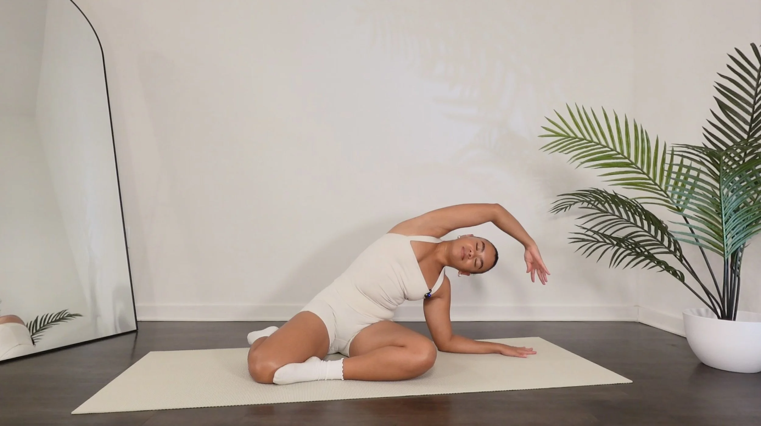 A woman practicing Pilates in a room with white walls and large green plants. She is sitting on a yoga mat, performing a mermaid stretch with one arm extended overhead and a peaceful expression.