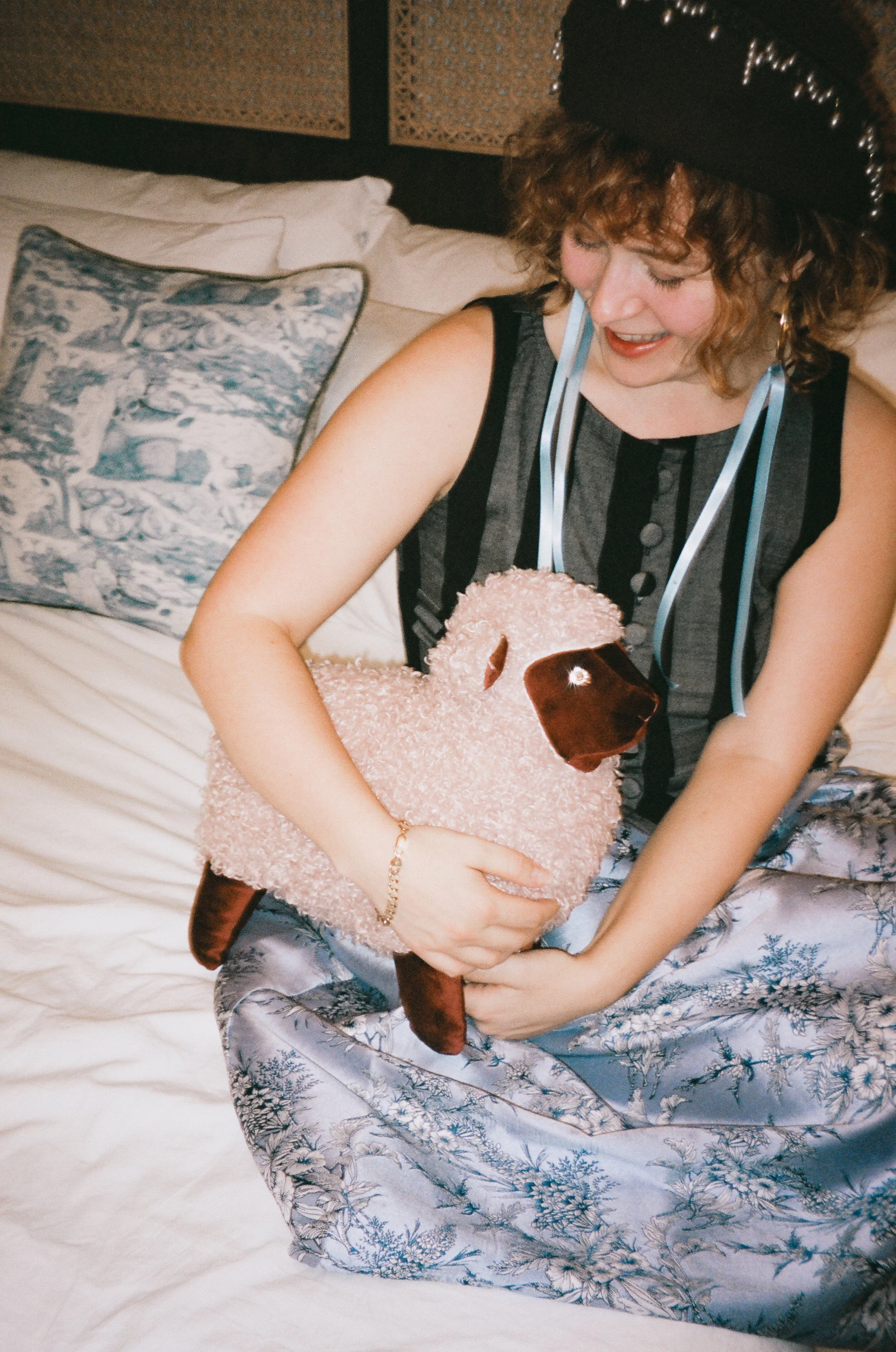 A woman with curly hair wearing a black hat with decorative beads, a sleeveless black and gray striped top, and a blue floral skirt, sitting on a bed holding a pink plush sheep toy and smiling.
