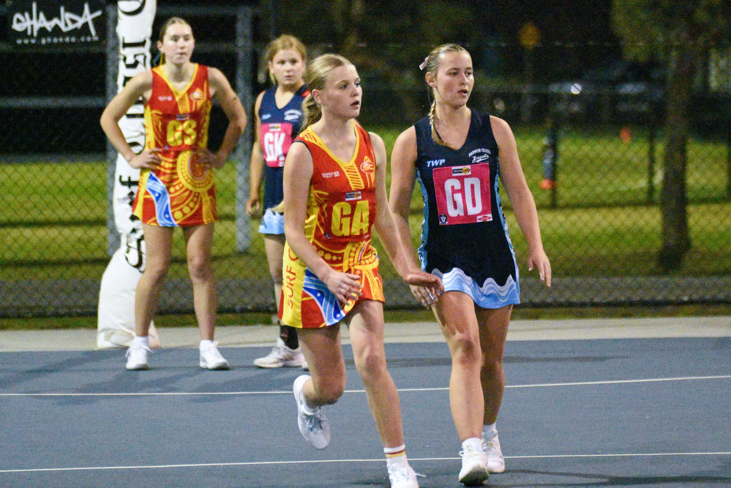 Four female netball players walking on a court during a game, wearing team uniforms with position labels, surrounded by a chain-link fence at night.