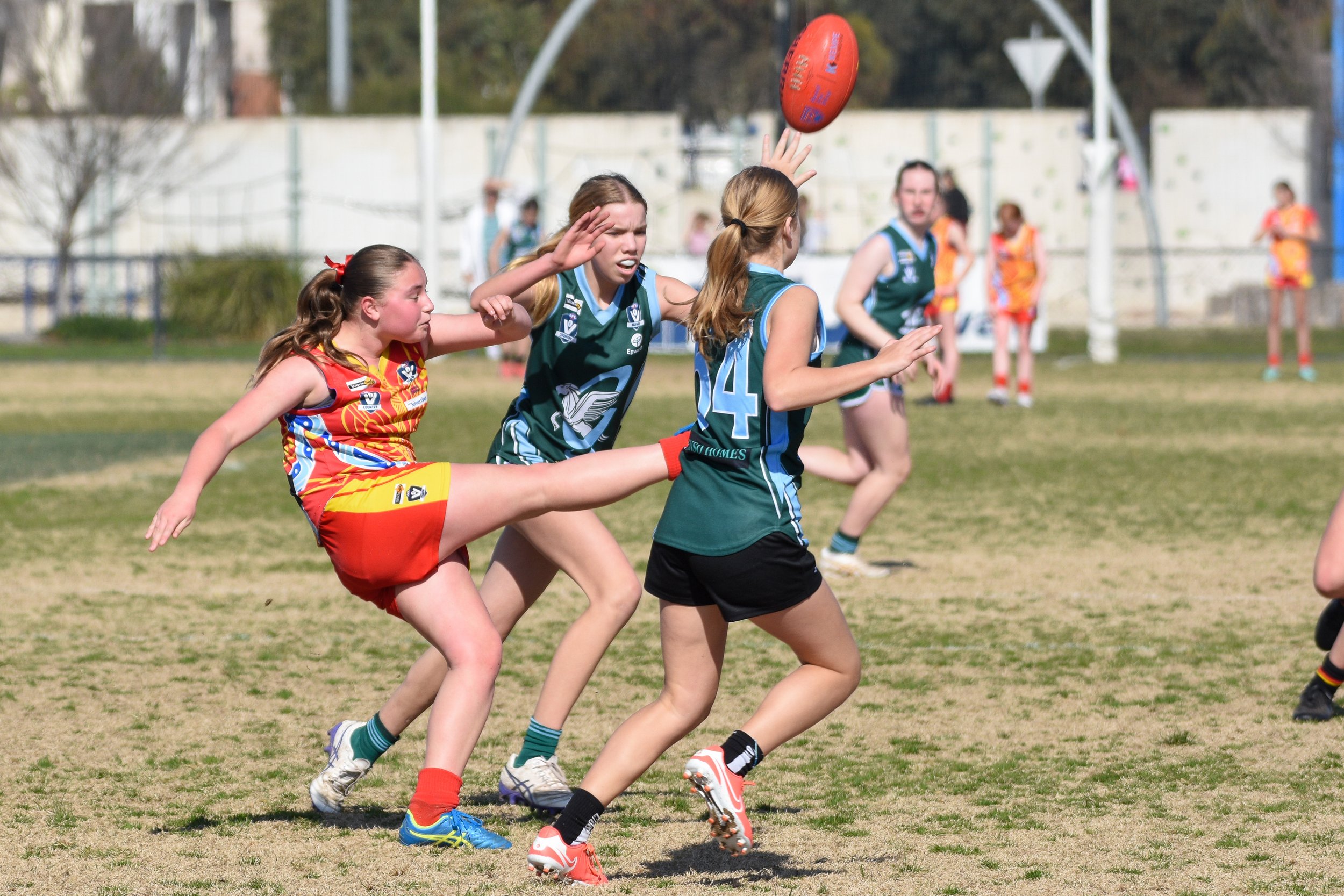 Girls playing Australian rules football on a grassy field during daytime, with one girl in a red and yellow uniform kicking the ball as others in green and blue uniforms attempt to catch it.