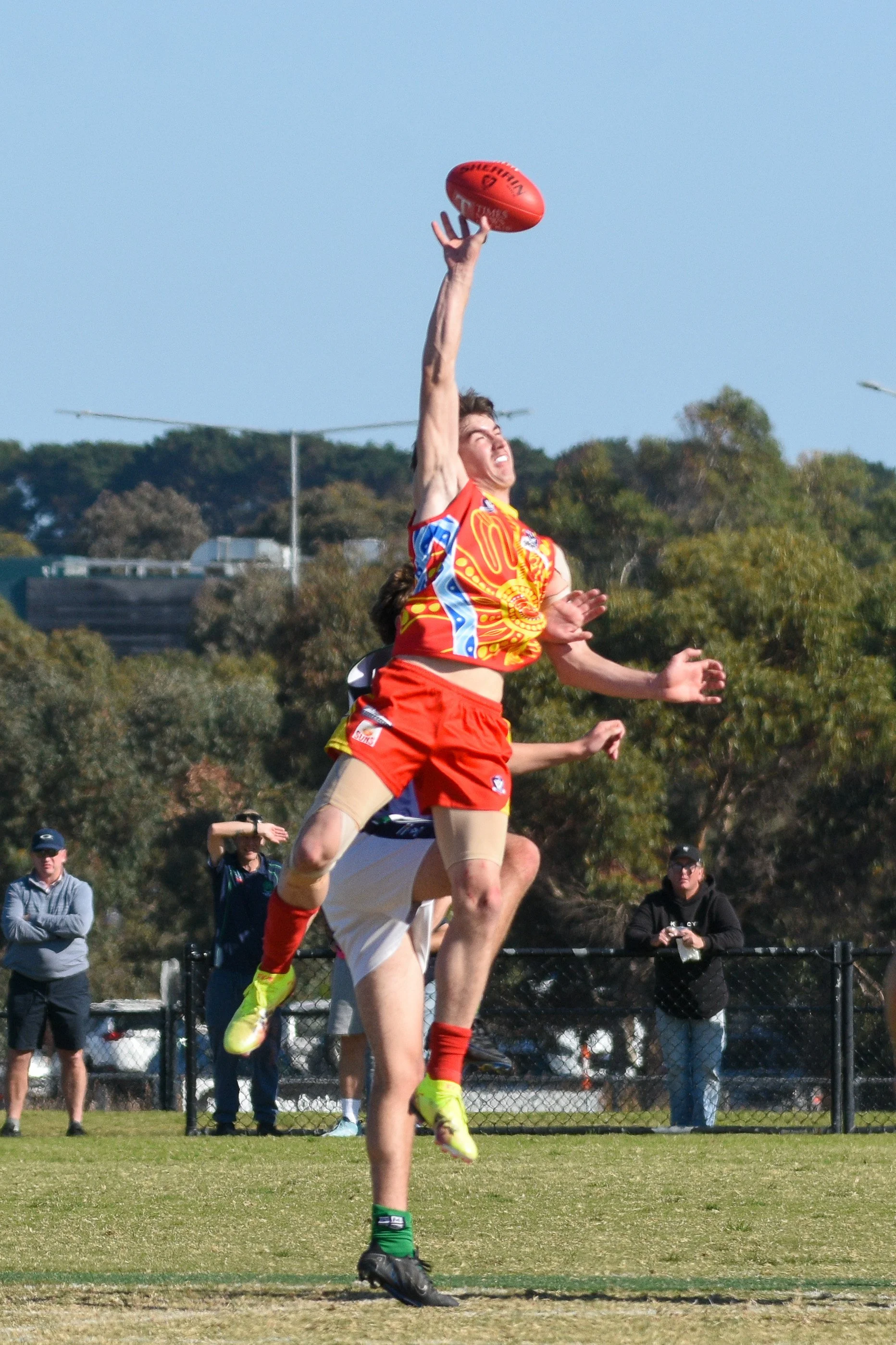 A athlete leaps to catch an Australian rules football while crowd members watch from behind a fence on a grassy field.