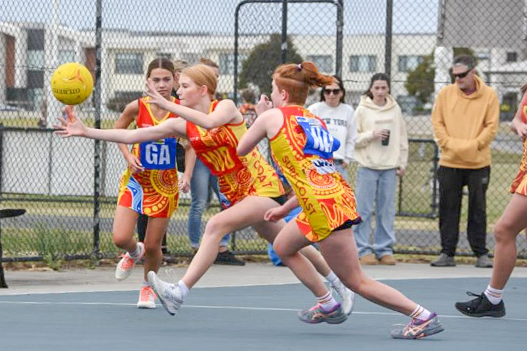 Girls in colorful uniforms playing netball on an outdoor court, with spectators in the background.