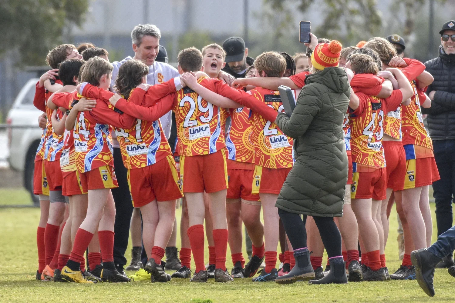 A group of young athletes in red and yellow sports uniforms huddle together in a circle on a soccer field, celebrating during daylight with some individuals smiling and hugging.