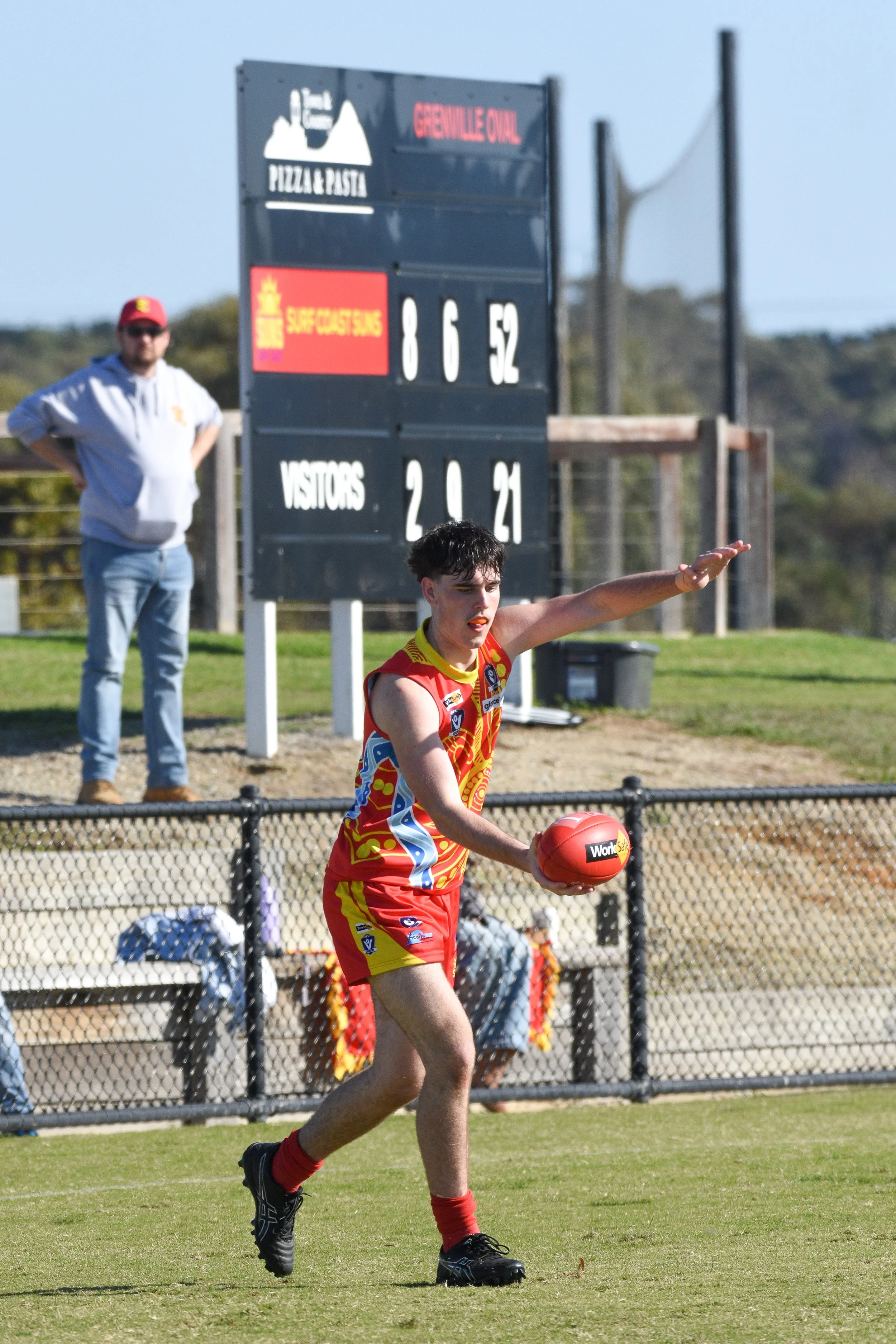 A young athlete in a red and yellow sports uniform holding an Australian football on a sports field, with a scoreboard and a man in casual clothing standing in the background.